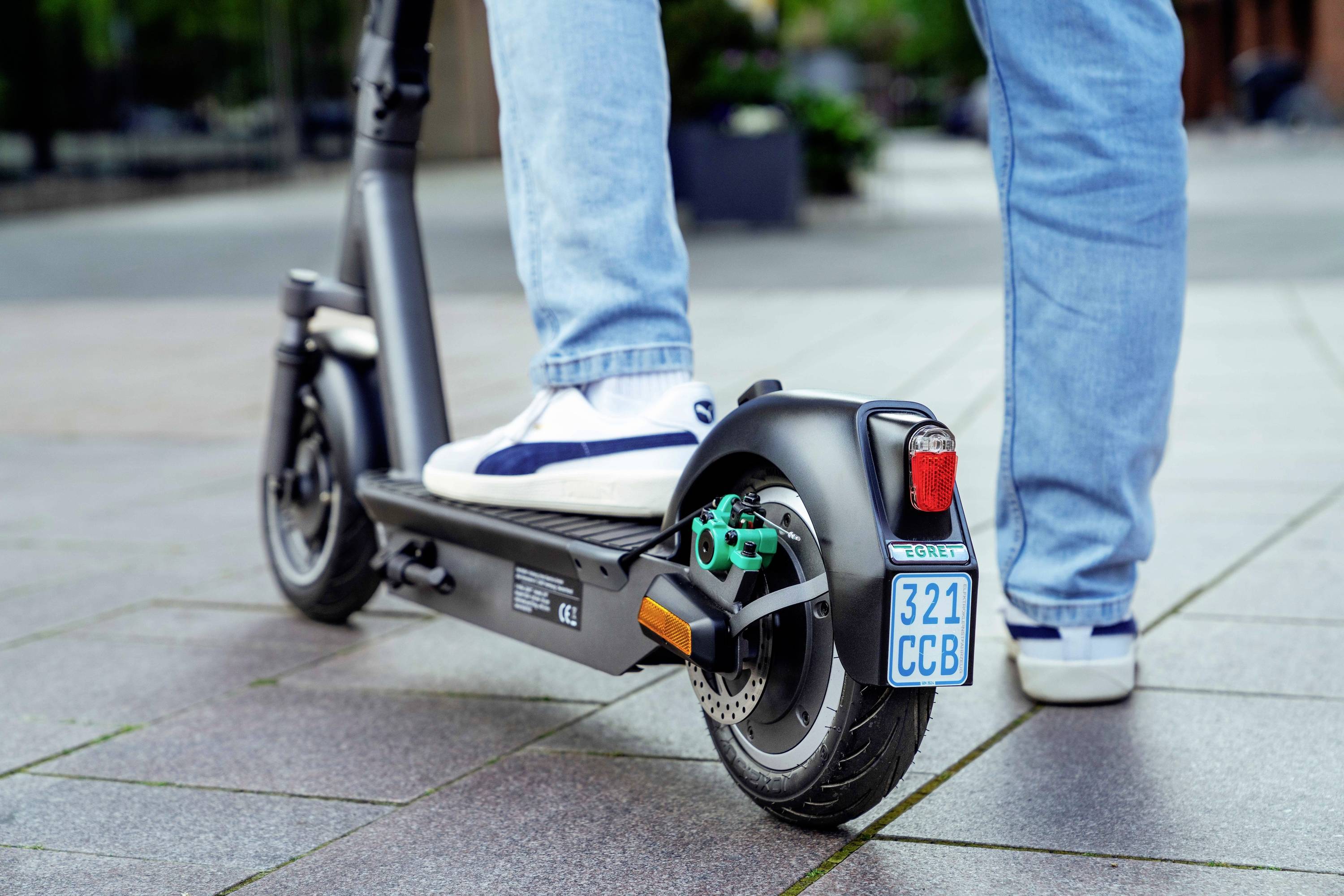 Eine Person fährt bei Tageslicht auf einem Bürgersteig einen Elektroroller. Der Roller hat eine kleine Nummernschildhalterung.