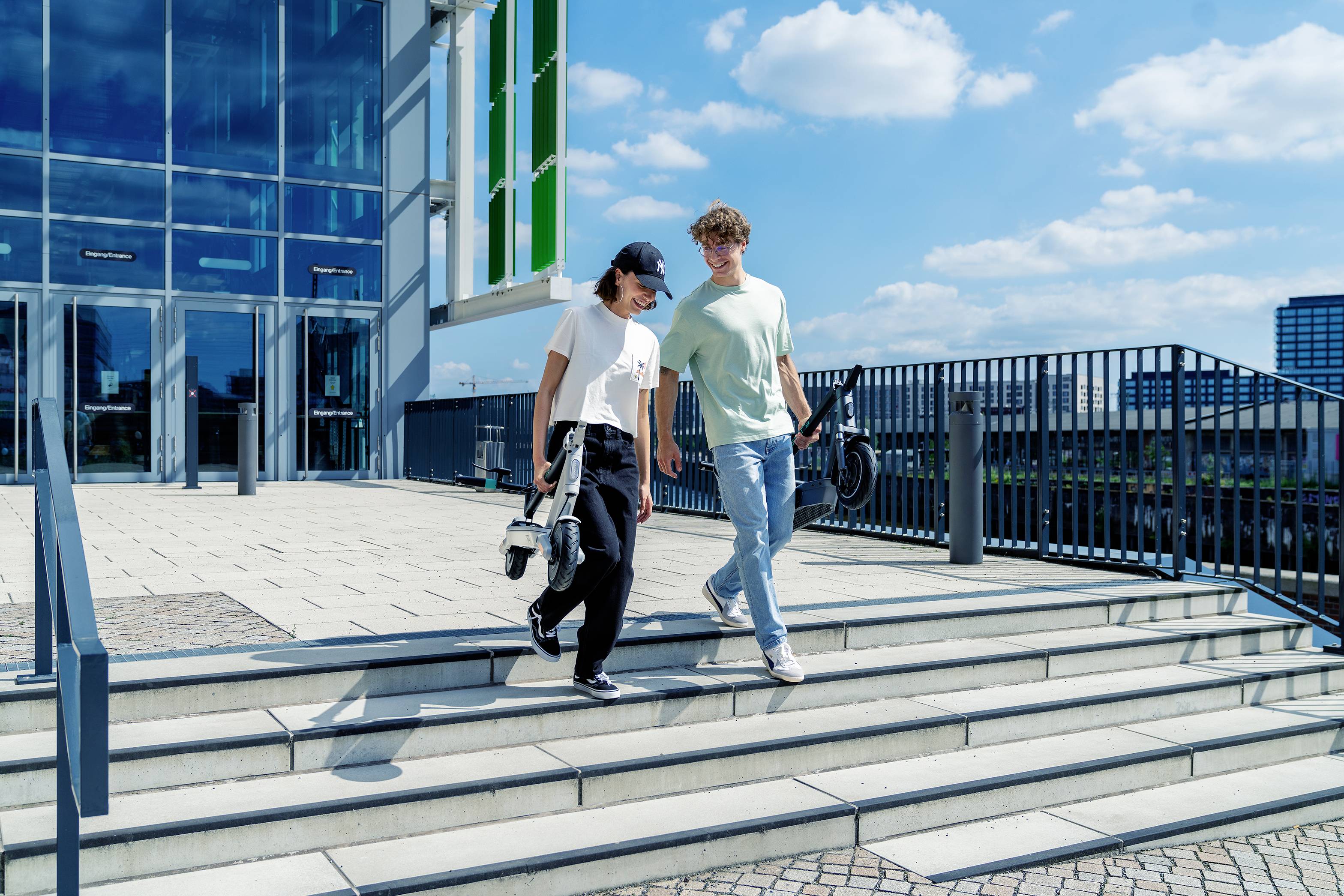 Zwei Personen gehen bei sonnigem Wetter eine Treppe hinunter, neben einem modernen Gebäude aus Glas.