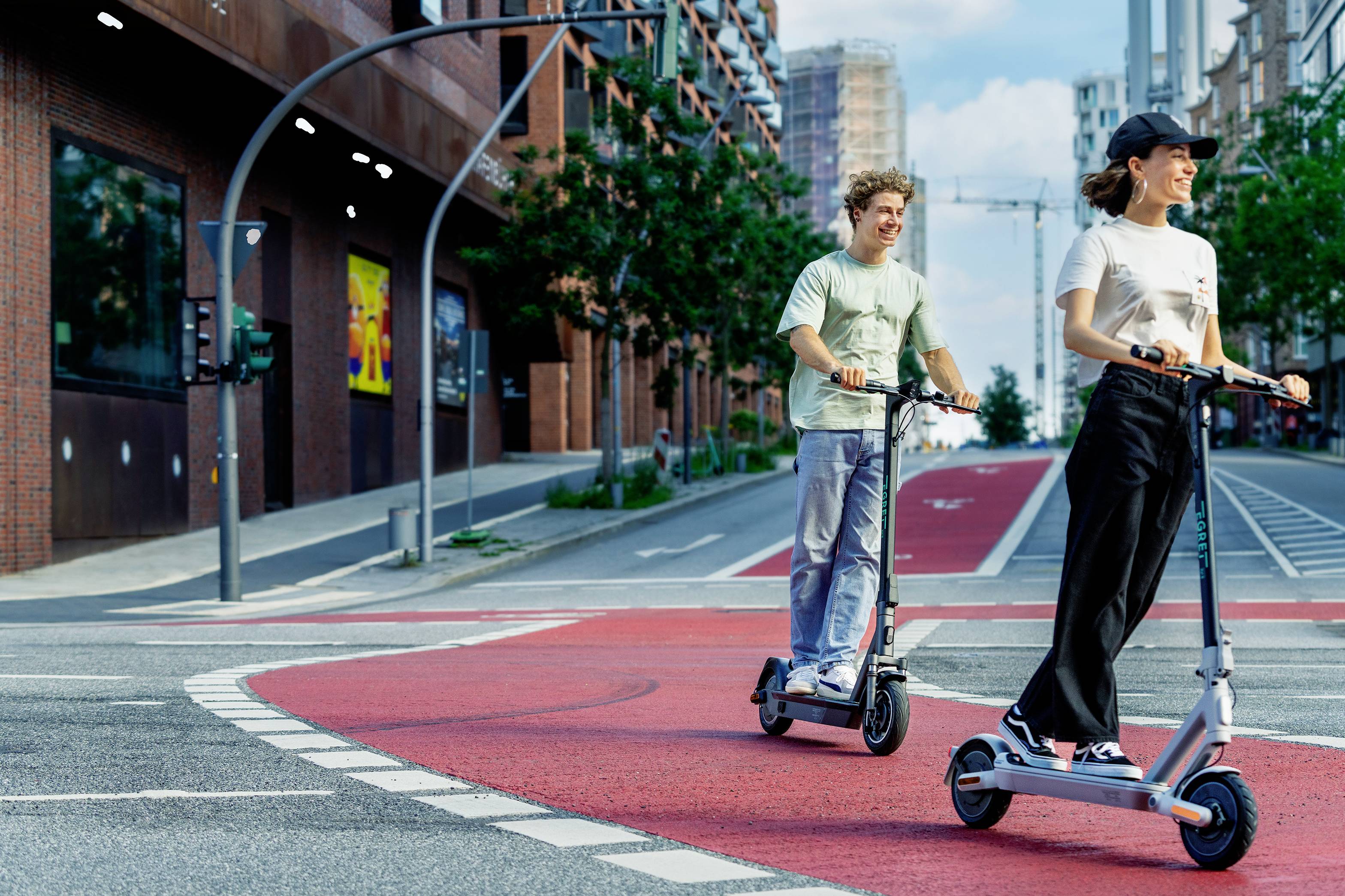 Zwei Personen fahren lachend auf E-Scootern auf einem roten Fahrradweg in einer urbanen Umgebung. Im Hintergrund sind Gebäude zu sehen.