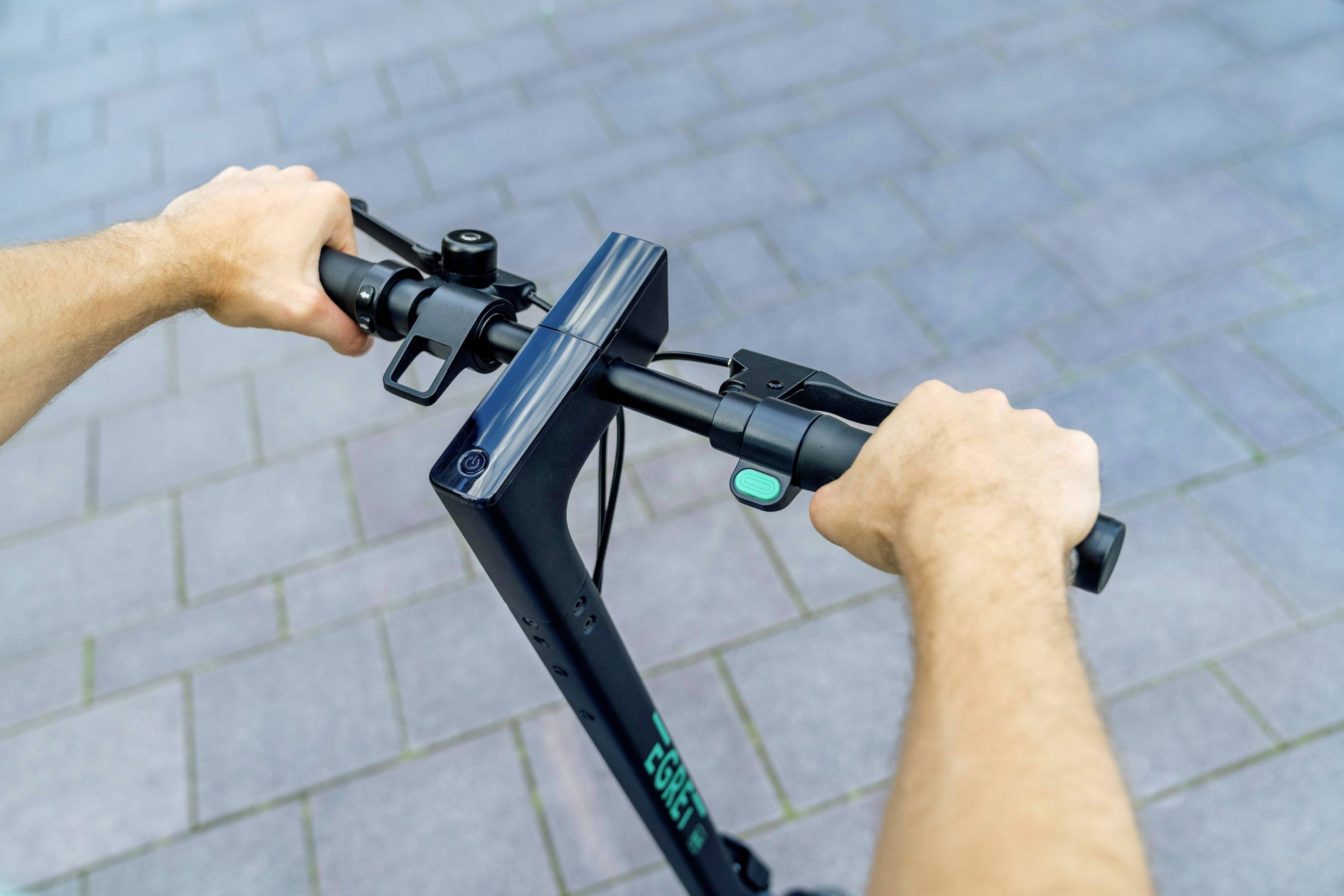 Zwei Hände halten die Lenker eines E-Scooters auf einer gepflasterten Straße. Der Daumen ruht auf einem grünen Knopf am Lenker.