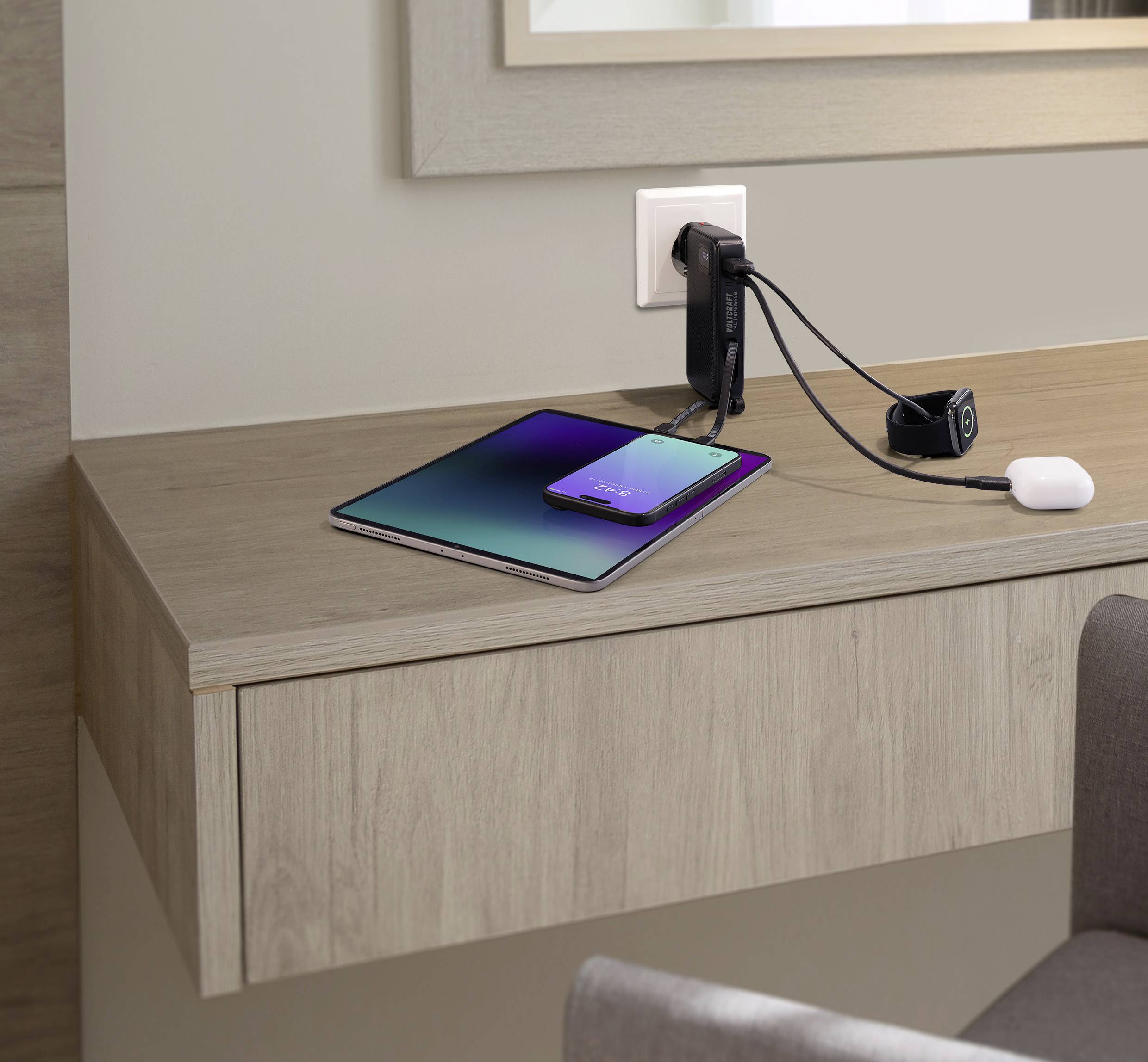 Ein Smartphone und Tablet liegen auf einem Holztisch, beide sind an einer Steckdose an der Wand angeschlossen, um geladen zu werden.