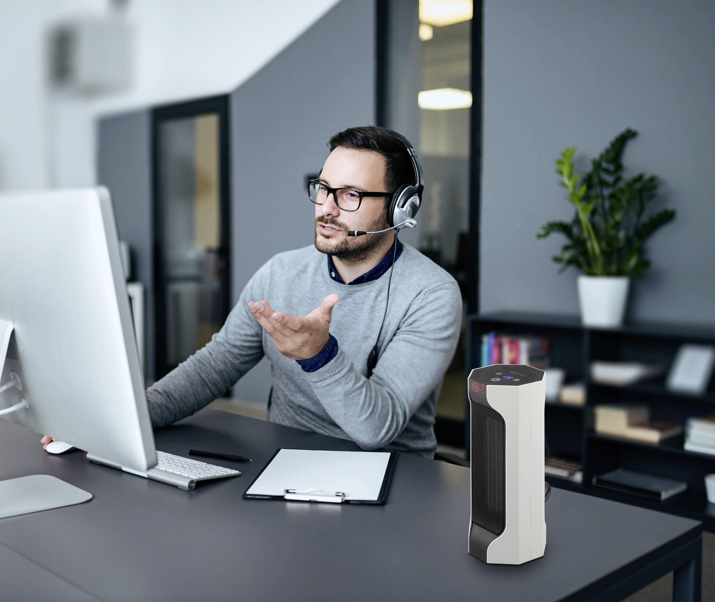 Ein Mann mit Kopfhörern arbeitet an einem Schreibtisch mit Computer und Notizblock. Im Hintergrund steht eine Pflanze.