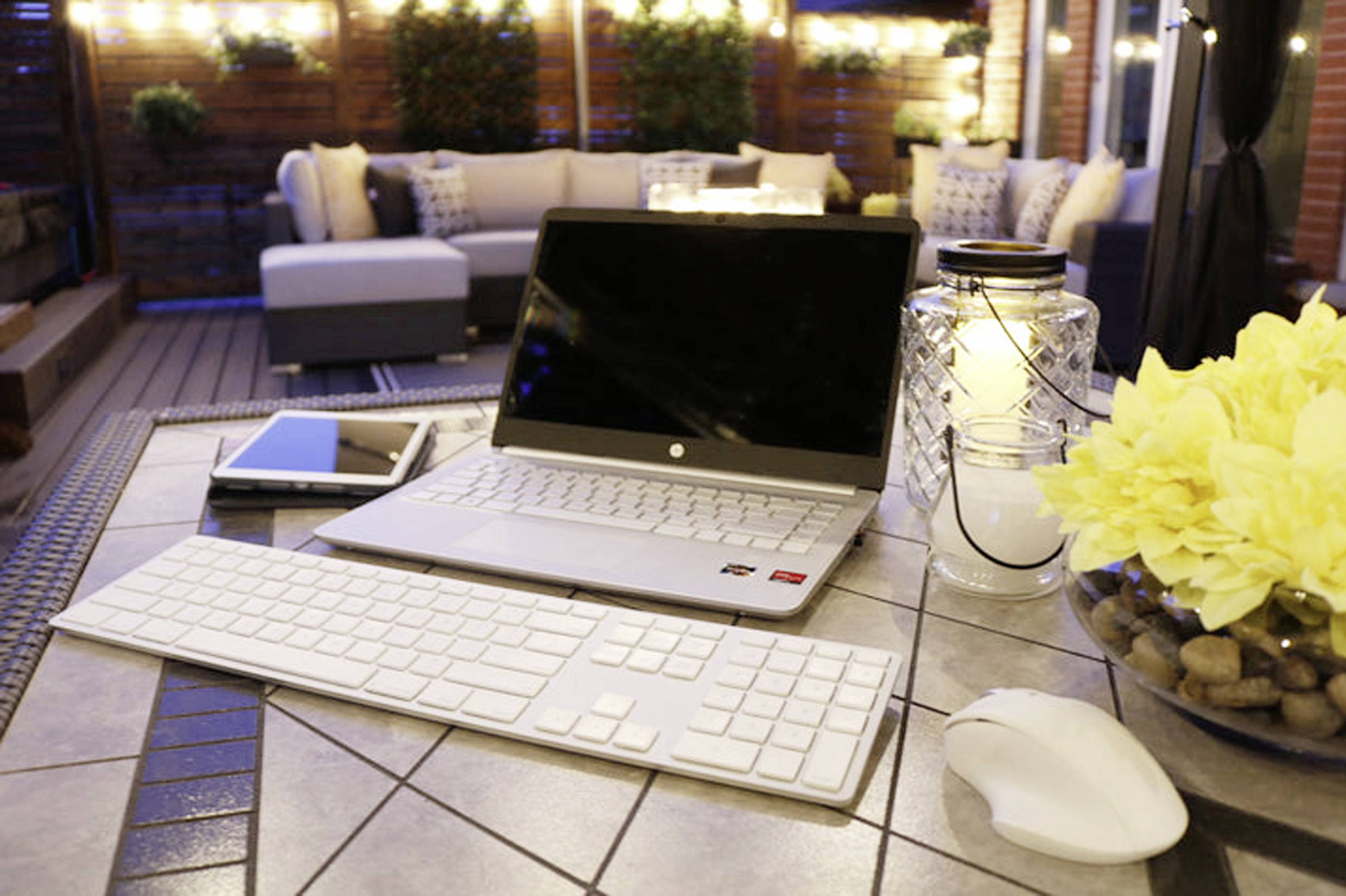 Laptop, Tablet, Tastatur und Maus auf einem Terrassentisch bei Abendbeleuchtung. Umgeben von Blumen und gemütlicher Sitzgruppe.