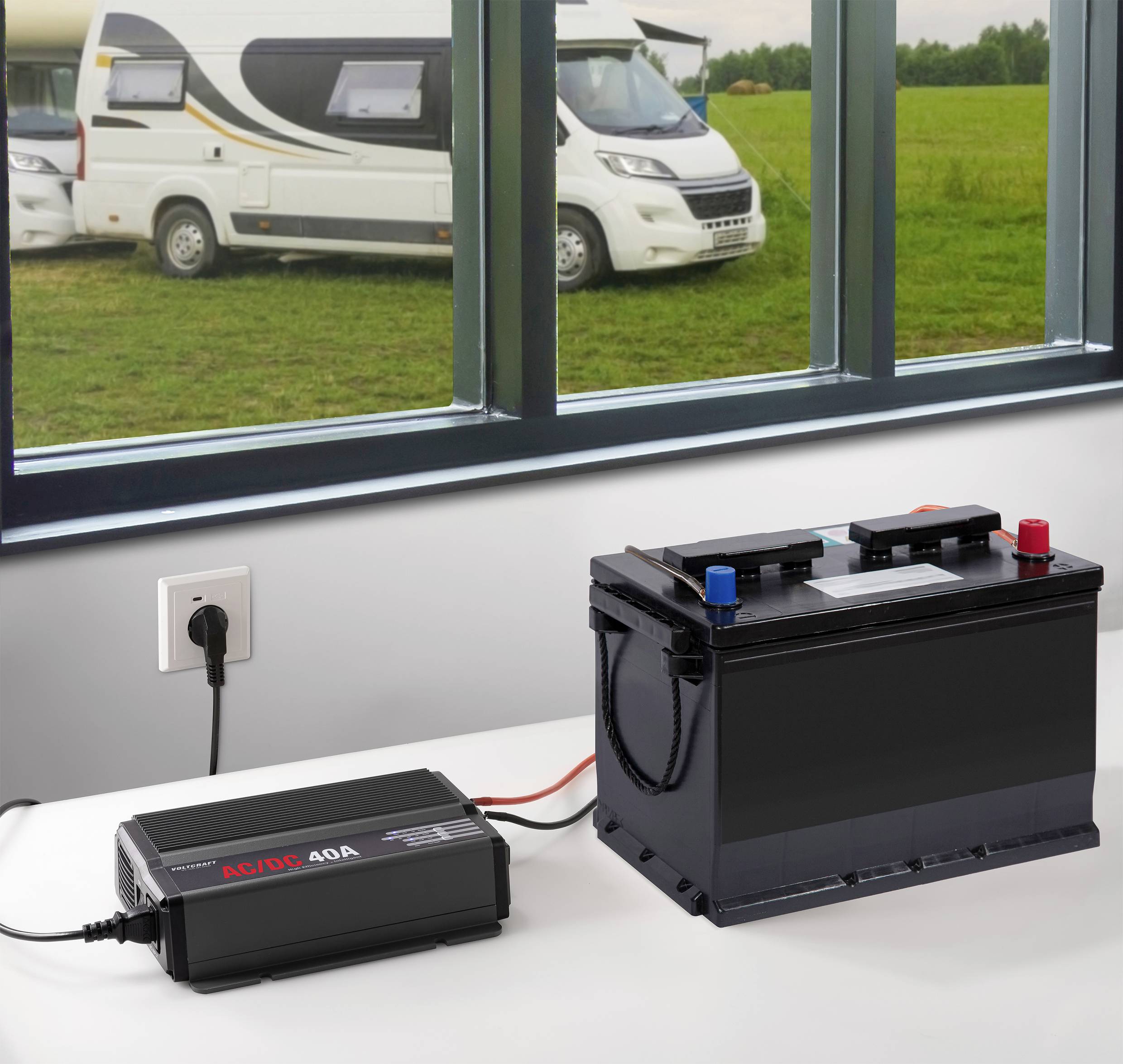 Ein Ladegerät ist mit einer Autobatterie auf einem Tisch verbunden. Im Hintergrund sind Wohnmobile und ein grünes Feld sichtbar.