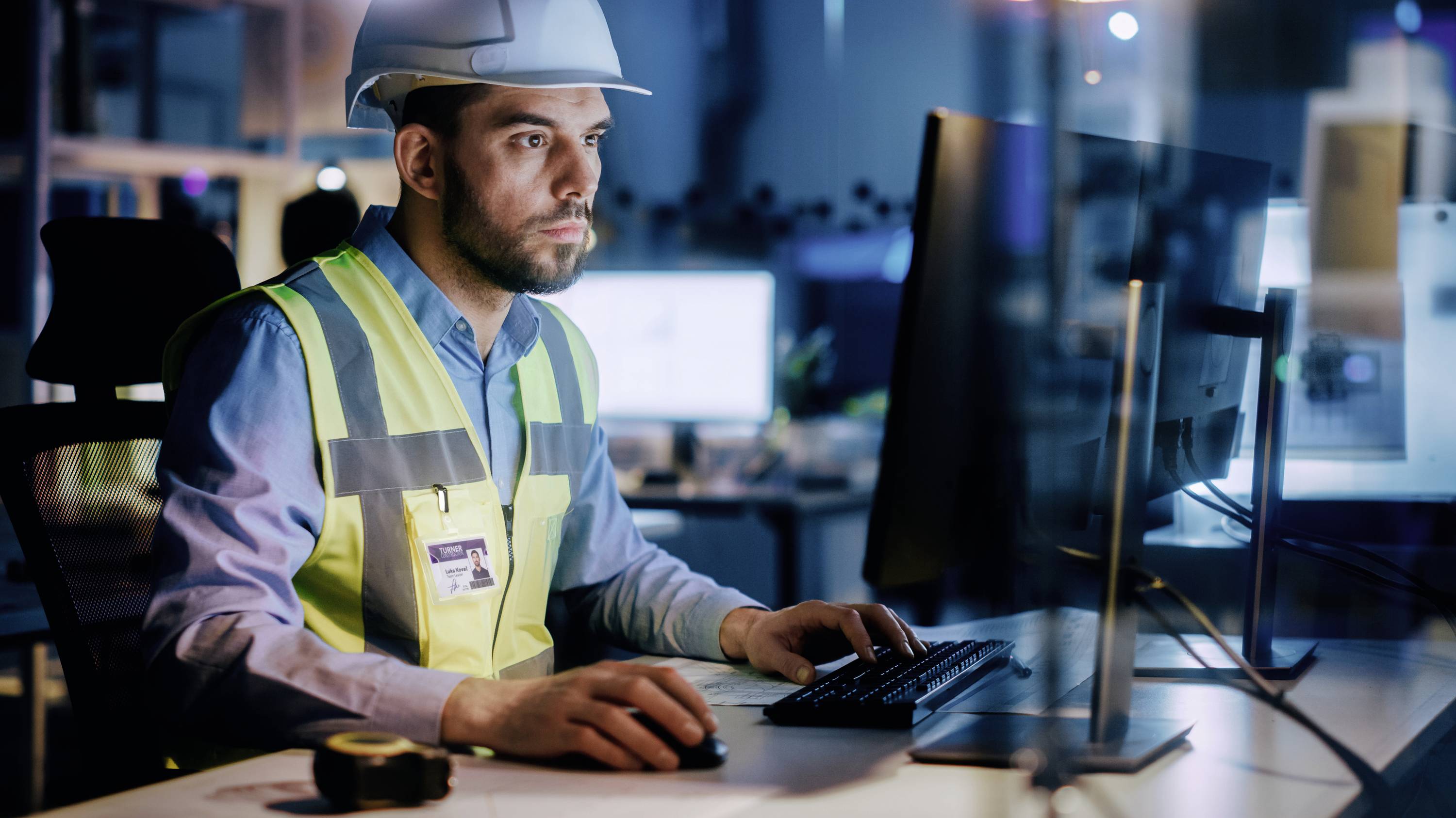 Ein Mann mit Helm und Sicherheitsweste arbeitet an einem Computerbildschirm in einem Büro, möglicherweise in einem Bauumfeld.