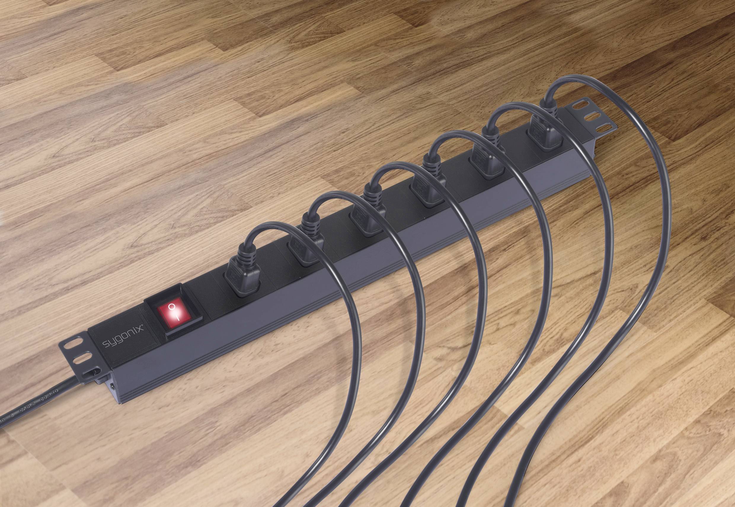 Ein Mehrfachsteckdose mit sechs eingesteckten schwarzen Kabeln liegt auf einem Holzfußboden. Der Ein-/Aus-Schalter ist rot beleuchtet.
