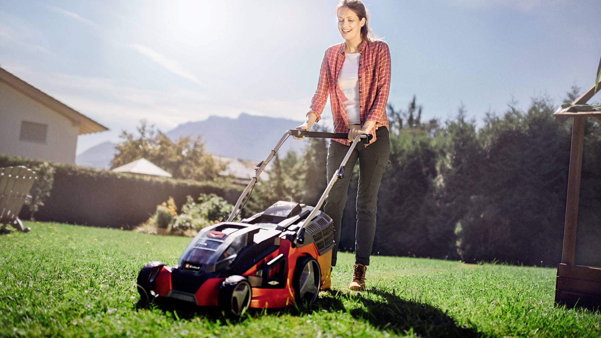 Eine Person mäht den Rasen mit einem roten Elektro-Rasenmäher an einem sonnigen Tag. Im Hintergrund sind Bäume und ein Haus zu sehen.