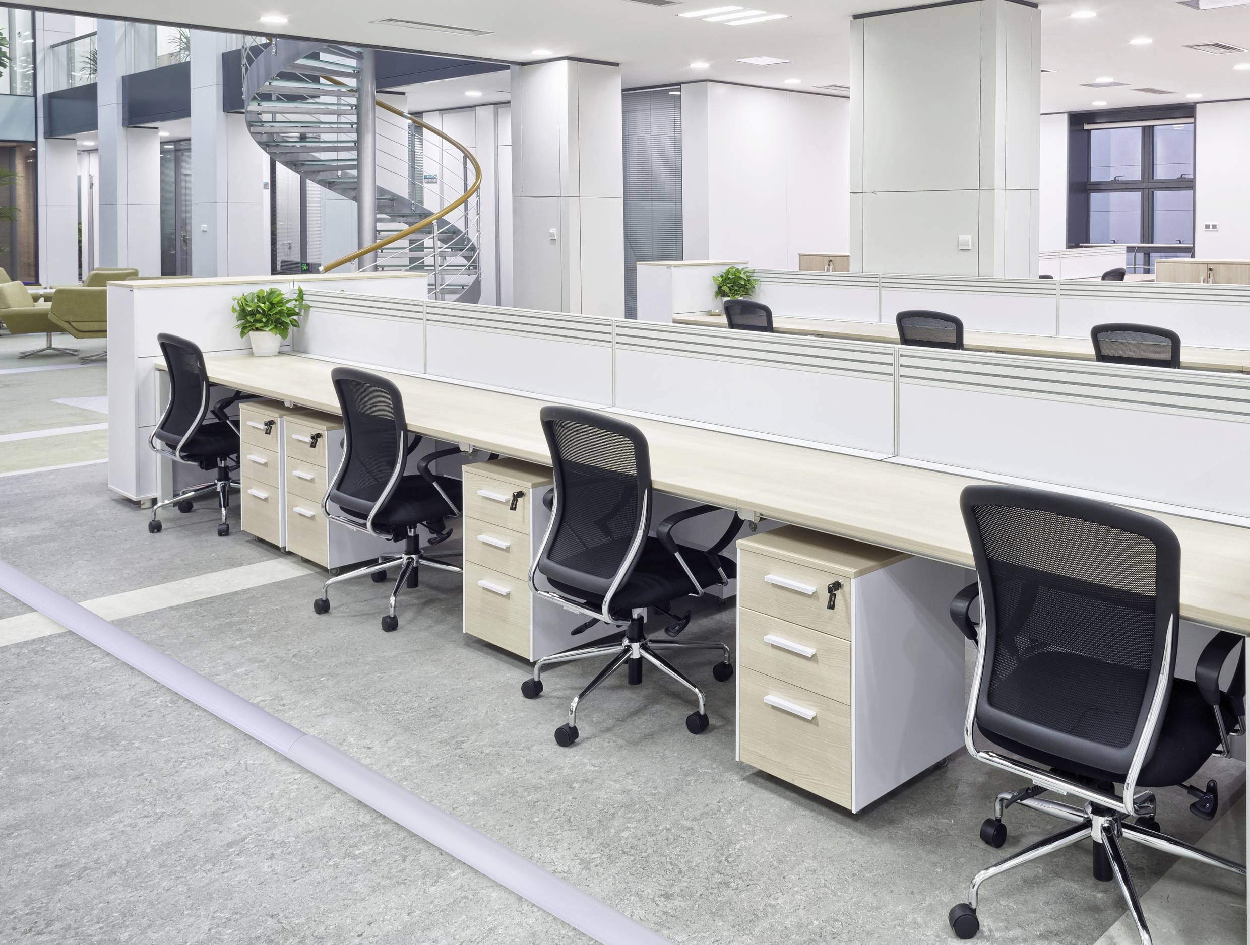 Großraumbüro mit mehreren leeren Schreibtischen und Bürostühlen, hell beleuchtet, mit modernen Möbeln und einer Wendeltreppe im Hintergrund.