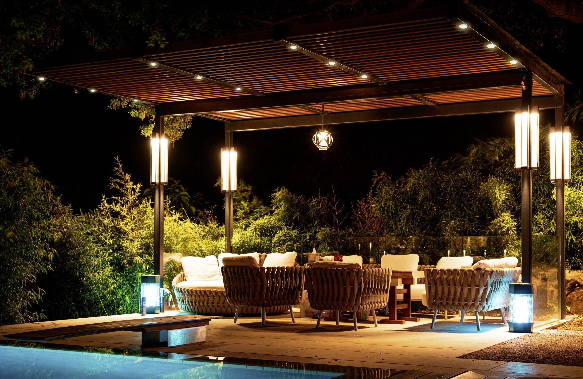 Eine gut beleuchtete Outdoor-Lounge bei Nacht mit gemütlichen Flechtmöbeln und einer Holzpergola, direkt neben einem Pool, umgeben von üppiger Vegetation.