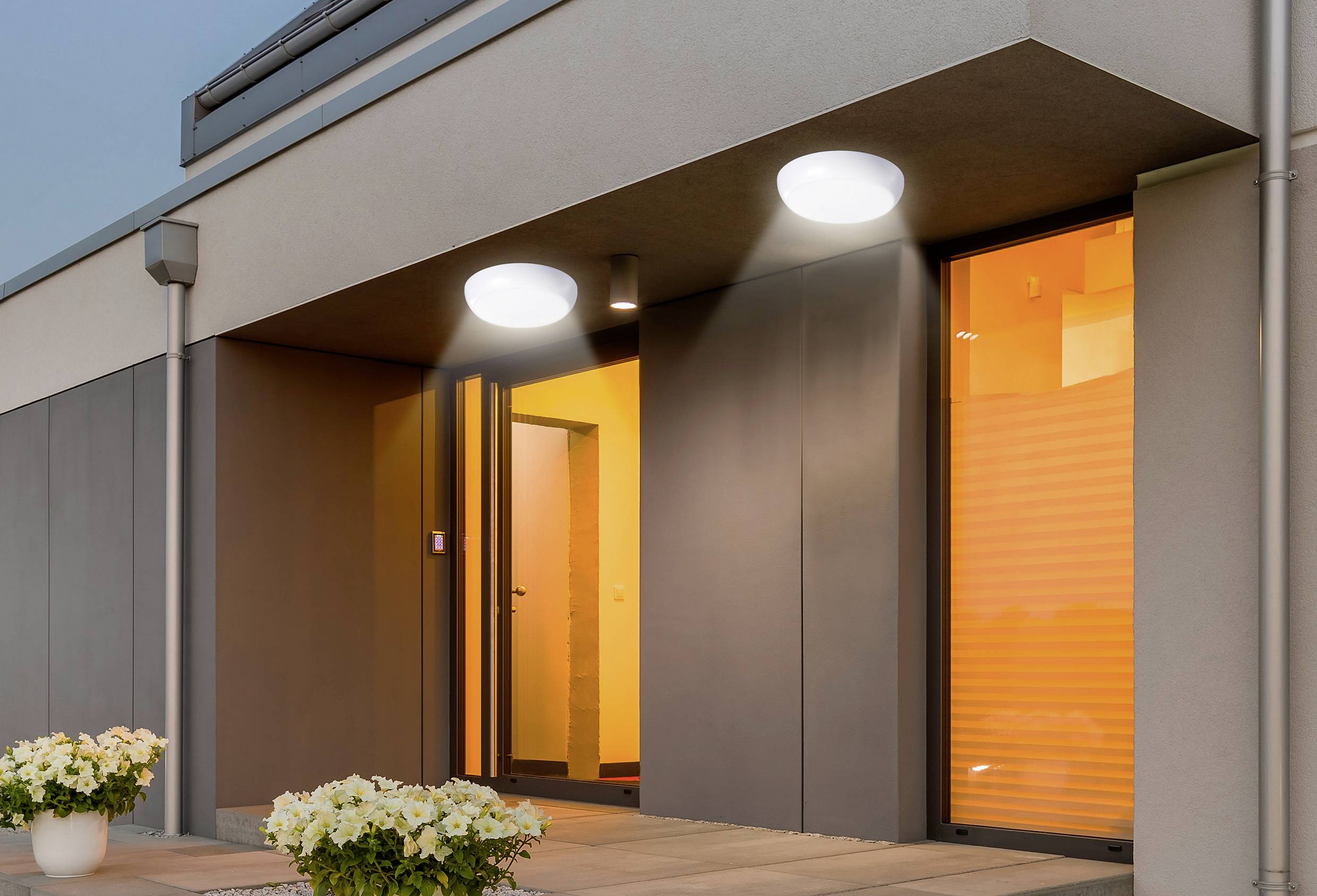 Ein moderner Gebäudeeingang mit beleuchteten Türöffnungen, Außenbeleuchtung, die Schatten wirft, und weißen Blumentöpfen auf der Terrasse.