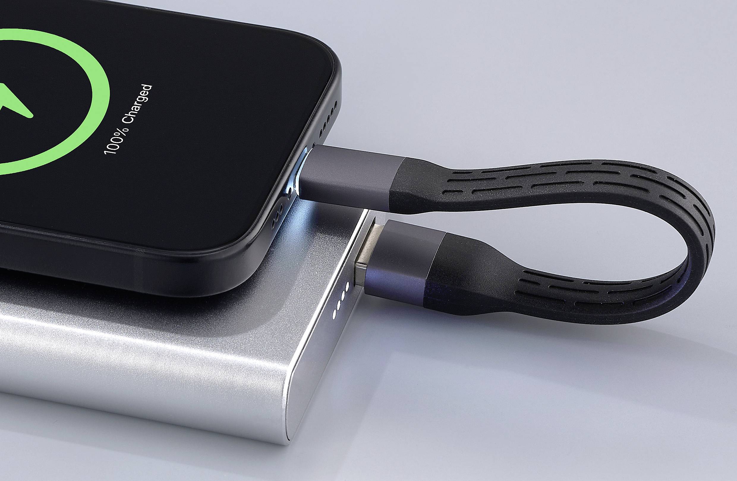 Ein Smartphone wird über ein schwarzes Kabel mit einer silbernen Powerbank aufgeladen. Auf dem Bildschirm wird '100% geladen' angezeigt.