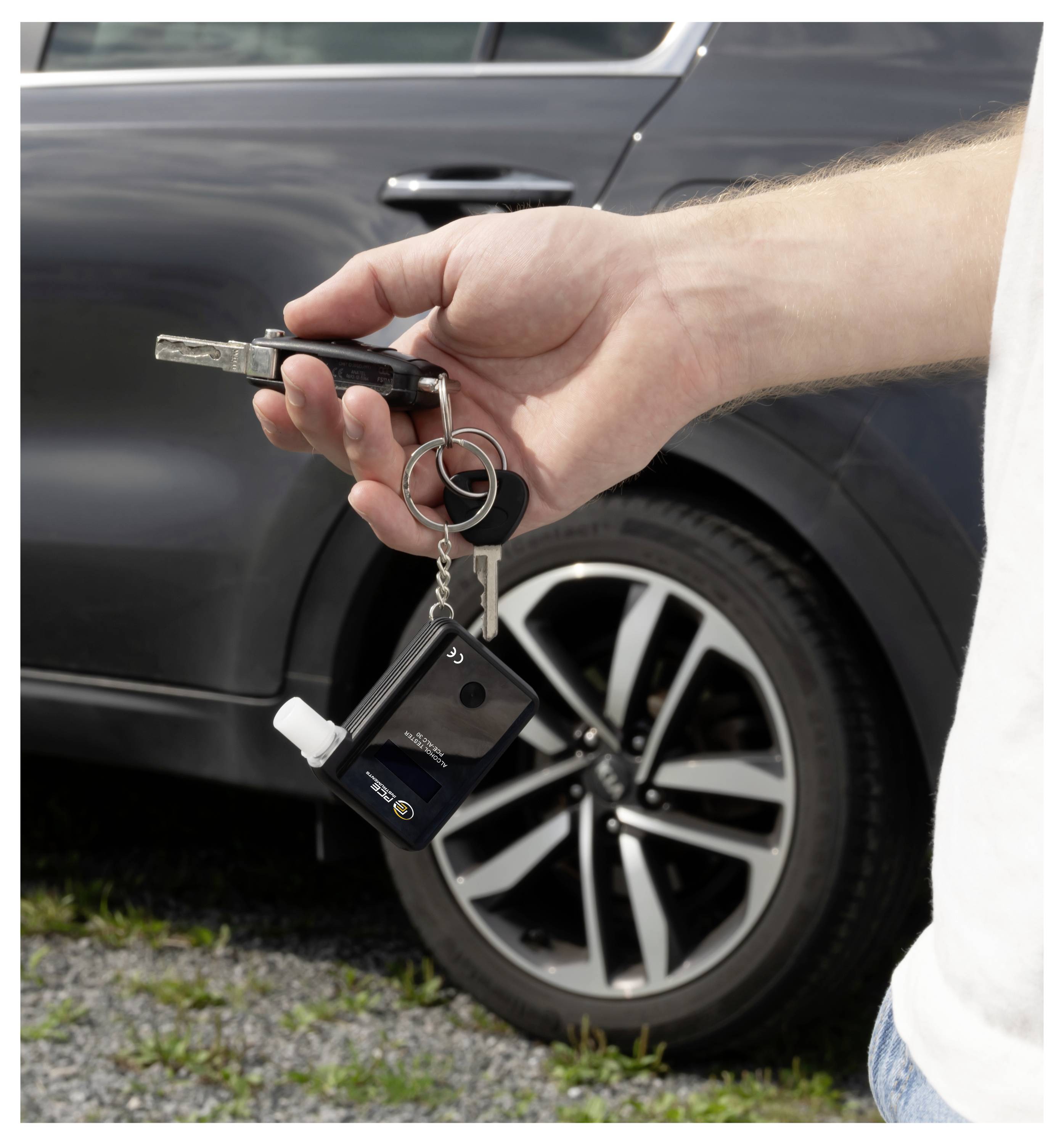 Hand hält Autoschlüssel und Schlüsselanhänger vor dem Rad eines dunklen Autos und deutet auf Fahrzeugzugang oder Eigentum hin.