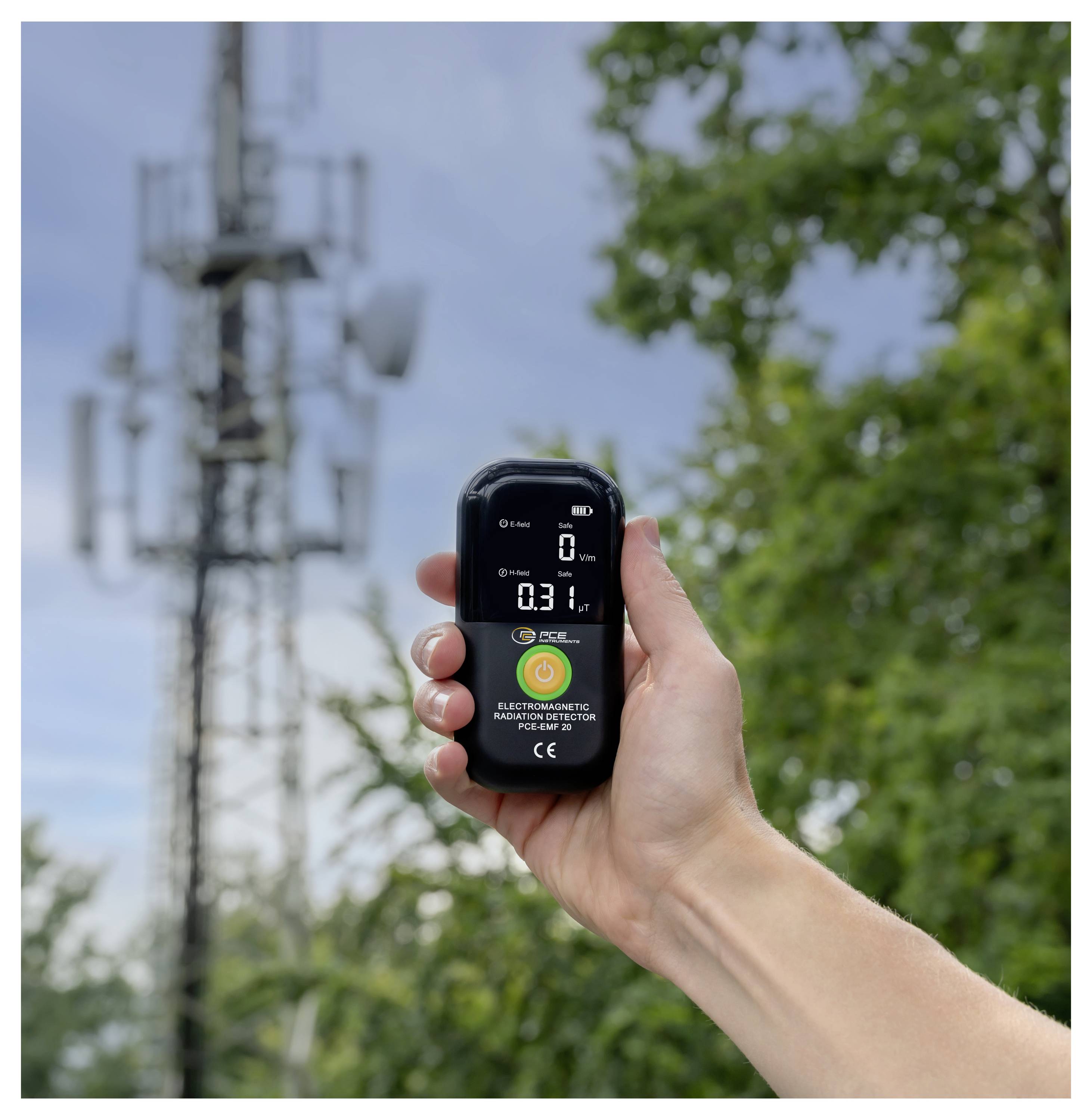 Hand hält einen digitalen Elektromagnetische-Strahlung-Detektor im Freien in der Nähe eines Mobilfunkmasts, wobei auf dem Bildschirm ein kleiner numerischer Wert angezeigt wird.