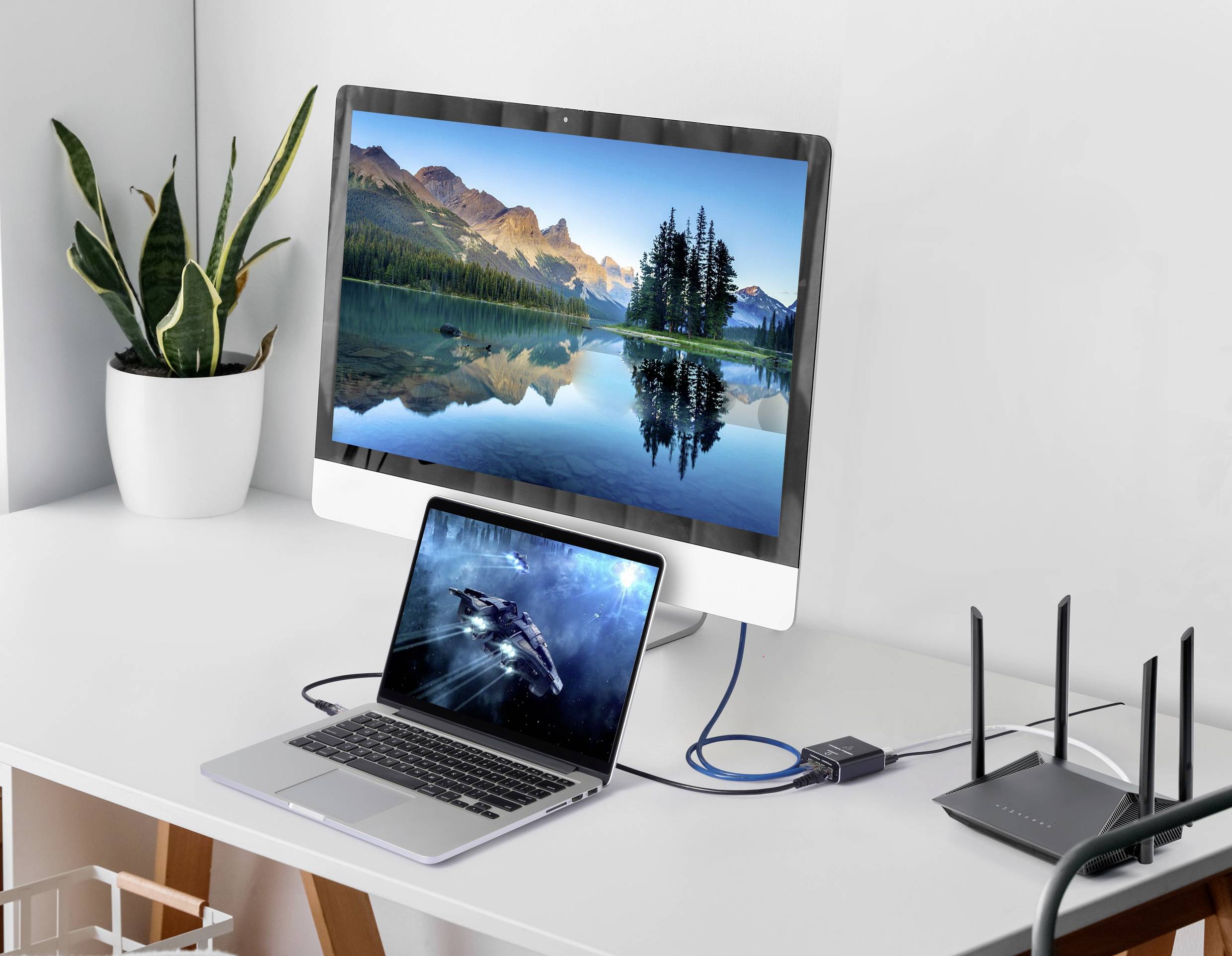 Ein Schreibtisch mit einem Monitor, der eine Landschaft zeigt, daneben ein Laptop und ein Router. Der Laptop ist über ein Kabel mit dem Monitor verbunden.