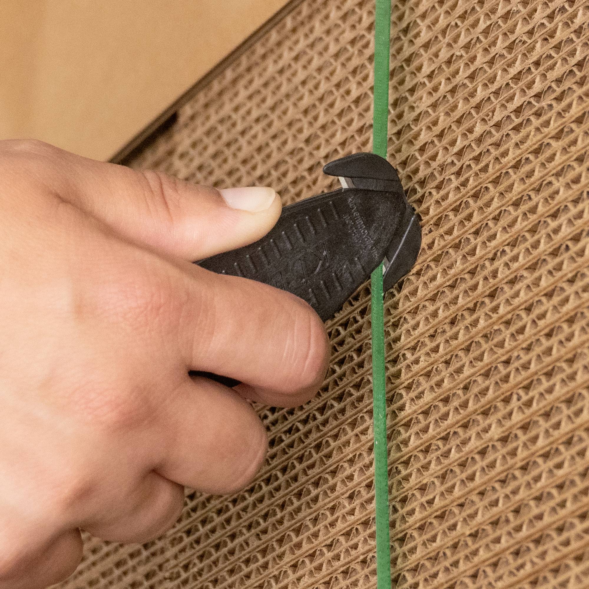 Nahaufnahme einer Hand, die mit einem Cutter-Messer eine grüne Plastikschnur auf einer braunen Wellpappe durchschneidet.