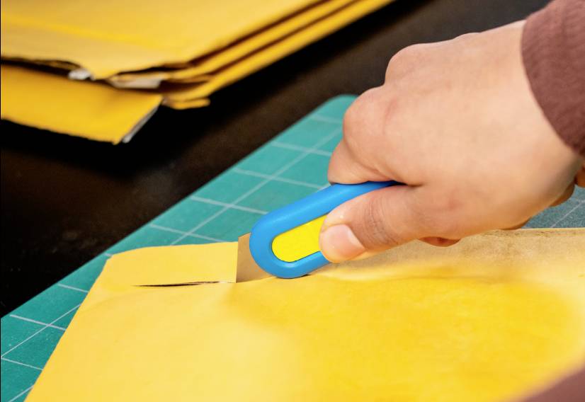 Eine Hand hält einen blauen Cuttermesser und schneidet einen braunen Umschlag auf einem grünen Schneidebrett.