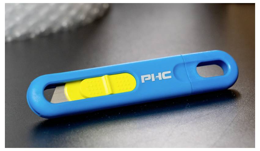 Ein blauer, rechteckiger Schieberegler mit gelbem Schalter, der auf einer grauen Oberfläche liegt. Darauf steht 'PHC'.