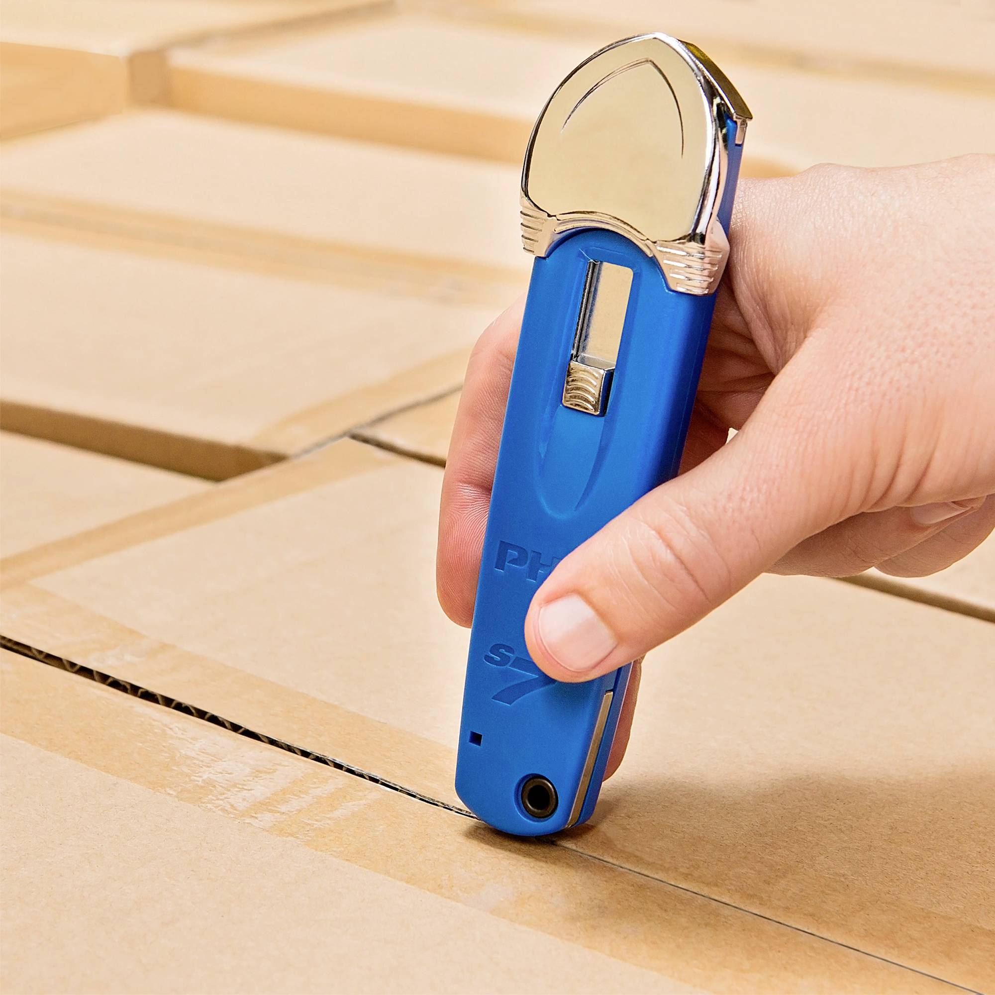 Eine Hand hält einen blauen Kartonschneider über einen Karton, bereit zum Schneiden. Der Karton ist gut gefaltet und geschlossen.