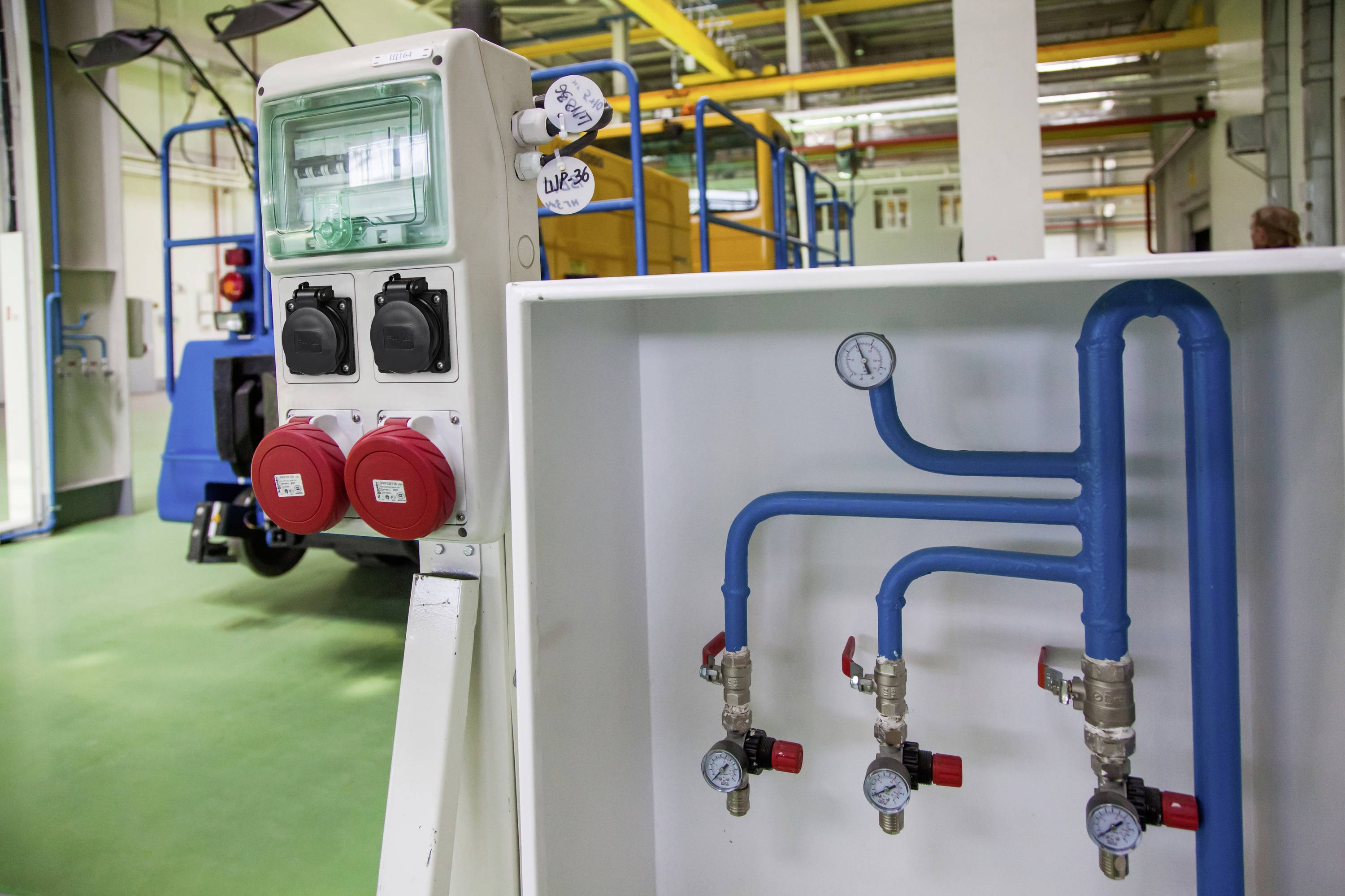 Industriehalle mit elektrischen Anschlüssen und blauen Rohrleitungen. Manometer zeigen Druck an. Gelbe und blaue Struktur im Hintergrund.