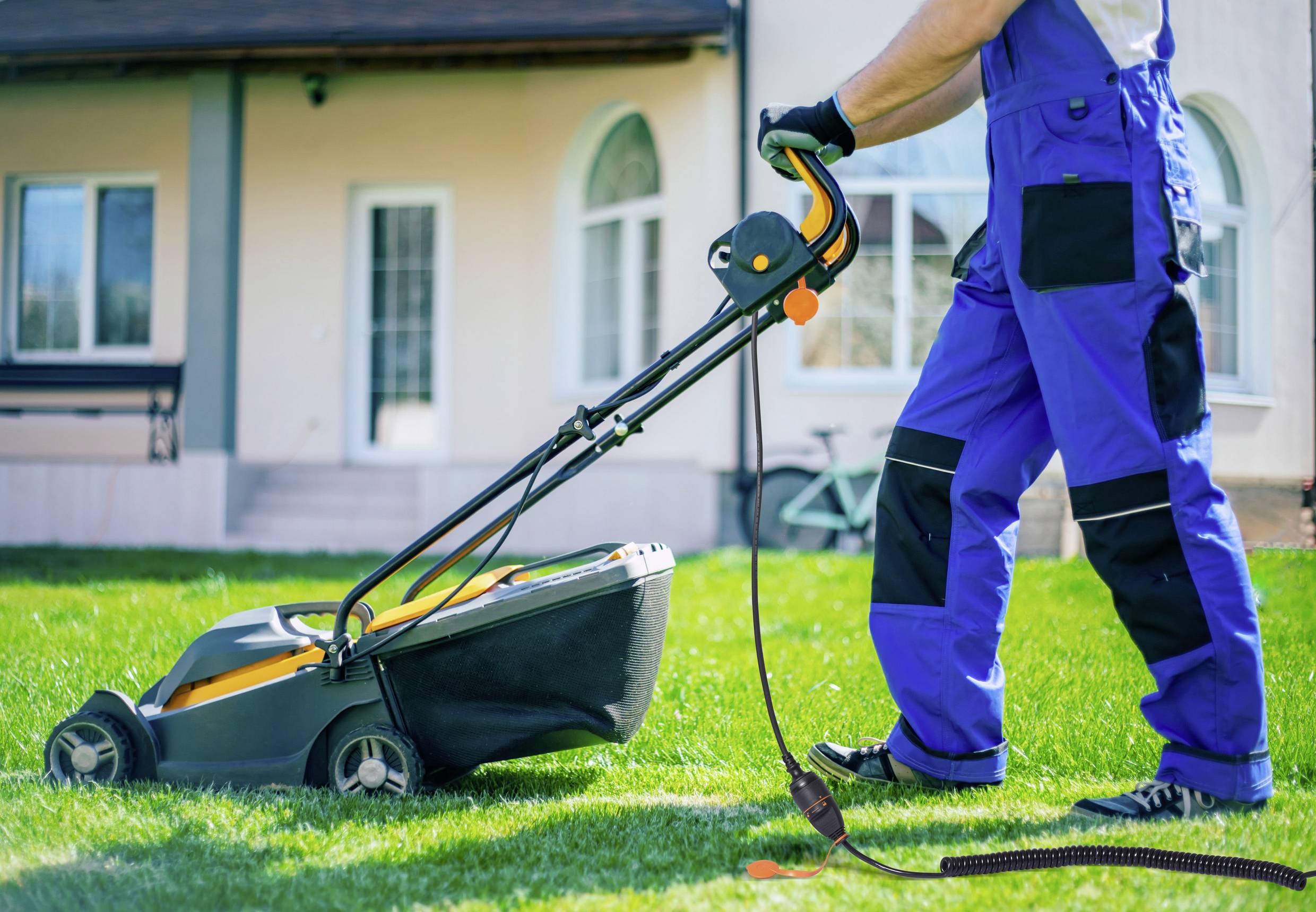 Person mäht Rasen mit elektrischem Rasenmäher; trägt blaue Arbeitskleidung. Haus im Hintergrund.
