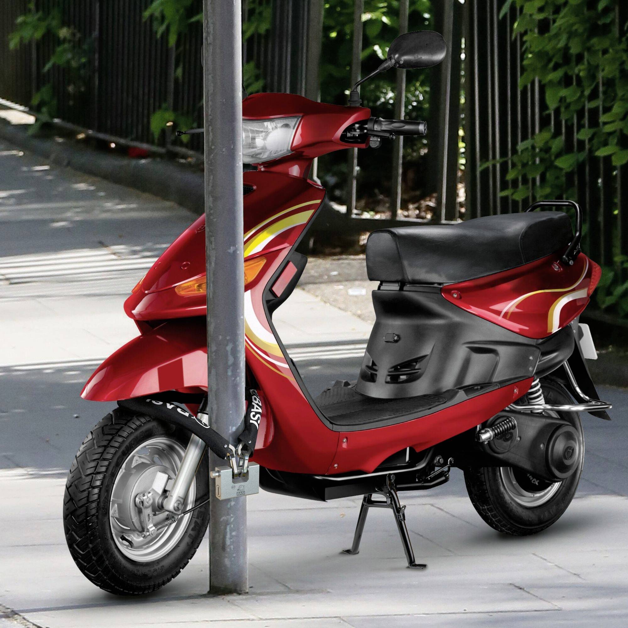 Ein roter Motorroller steht an einen hohen Metallpfosten gelehnt auf einem Bürgersteig neben einem Zaun mit grünen Blättern im Hintergrund.