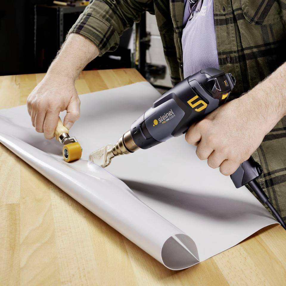 Ein Mann verwendet ein Heißluftgebläse und eine Rolle, um Folie auf einem Tisch zu verkleben.