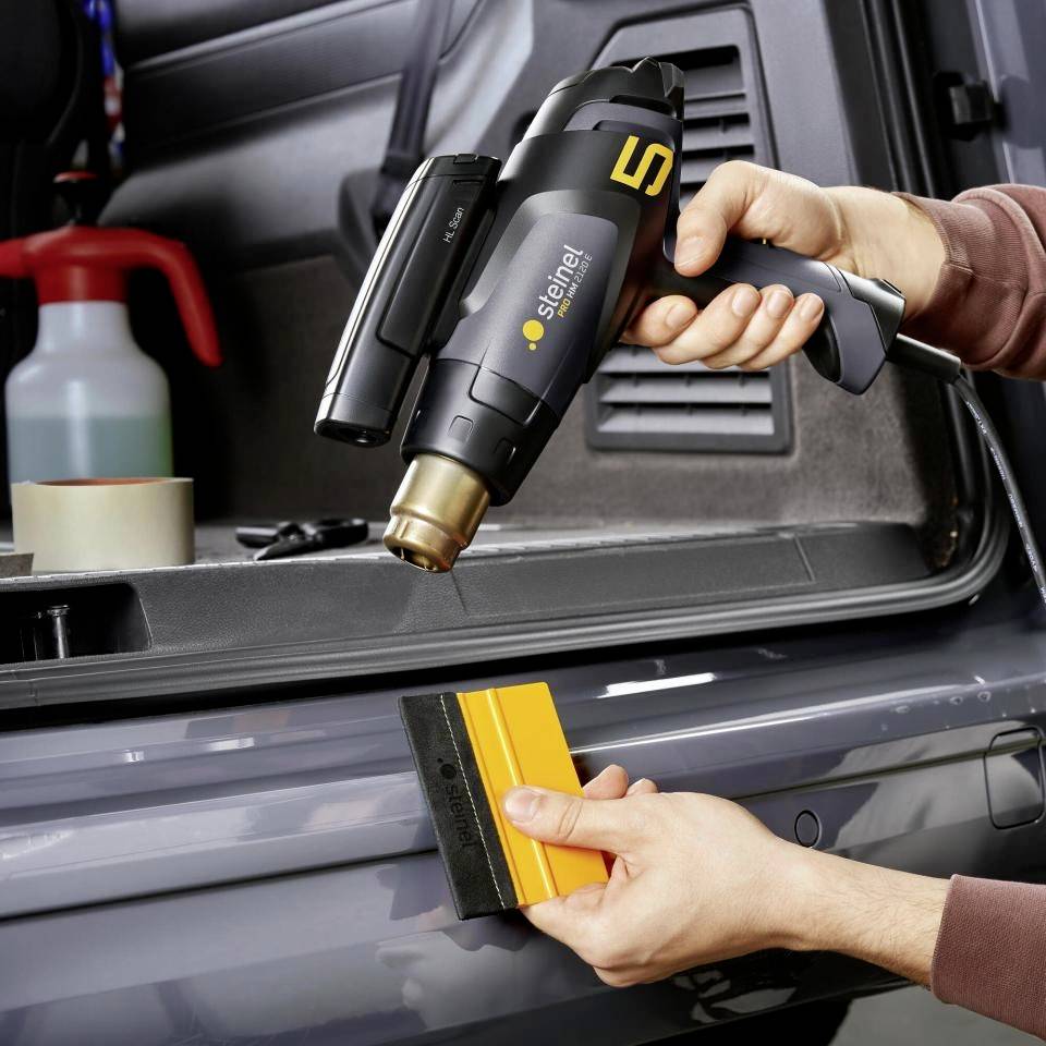 Eine Person hält ein schwarzes Heißluftgebläse und einen gelben Spachtel am Heck eines Autos. Im Hintergrund sind Werkzeuge zu sehen.