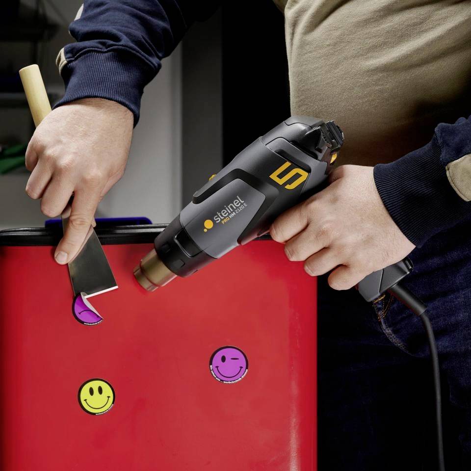 Ein Handwerker erhitzt mit einem Heißluftgebläse einen roten Kunststoff, um ihn zu formen. Ein Smiley-Aufkleber ist sichtbar.