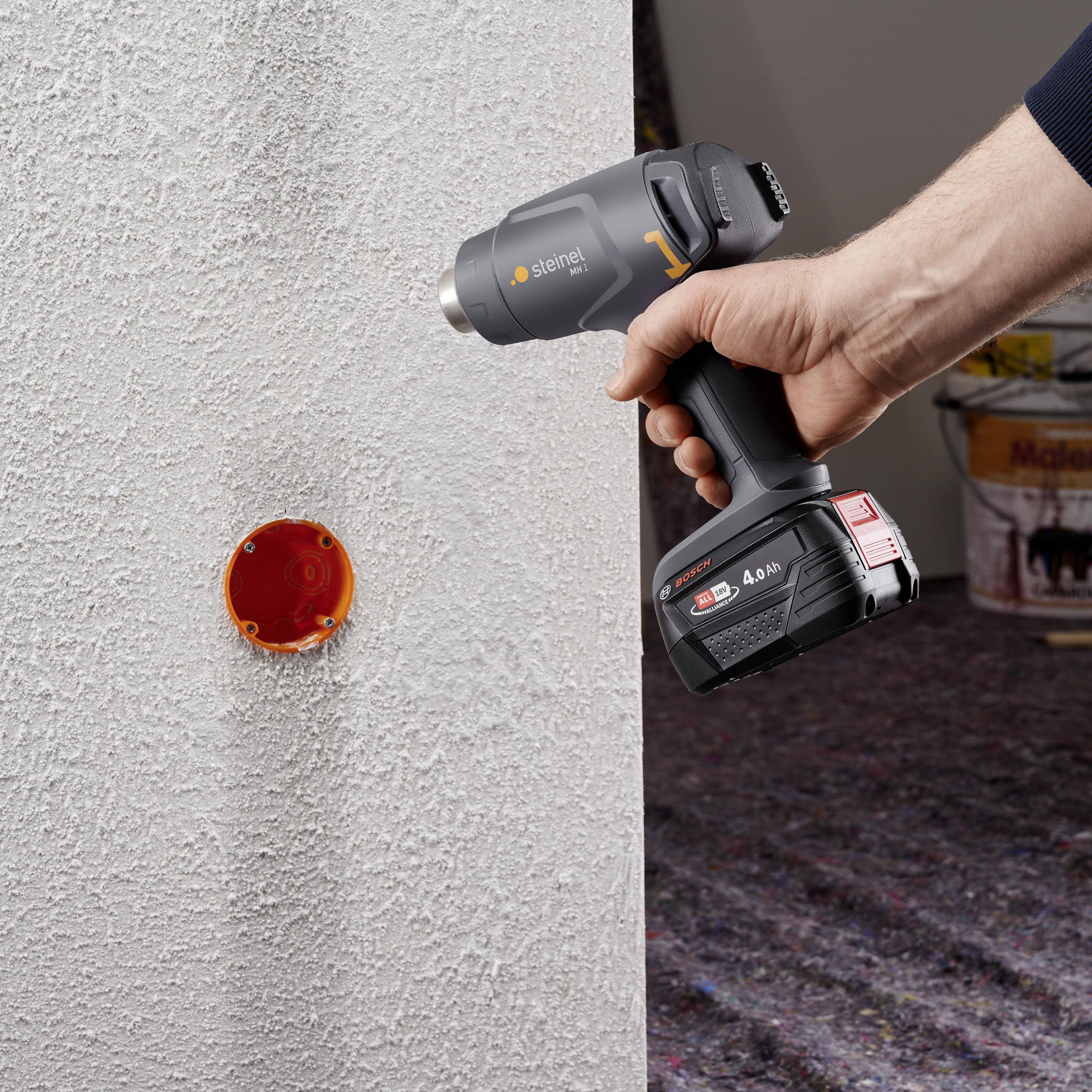 Eine Person hält ein graues Elektrowerkzeug an eine weiße Wand, wobei ein orangefarbener Kunststoff-Zylinder zu sehen ist.