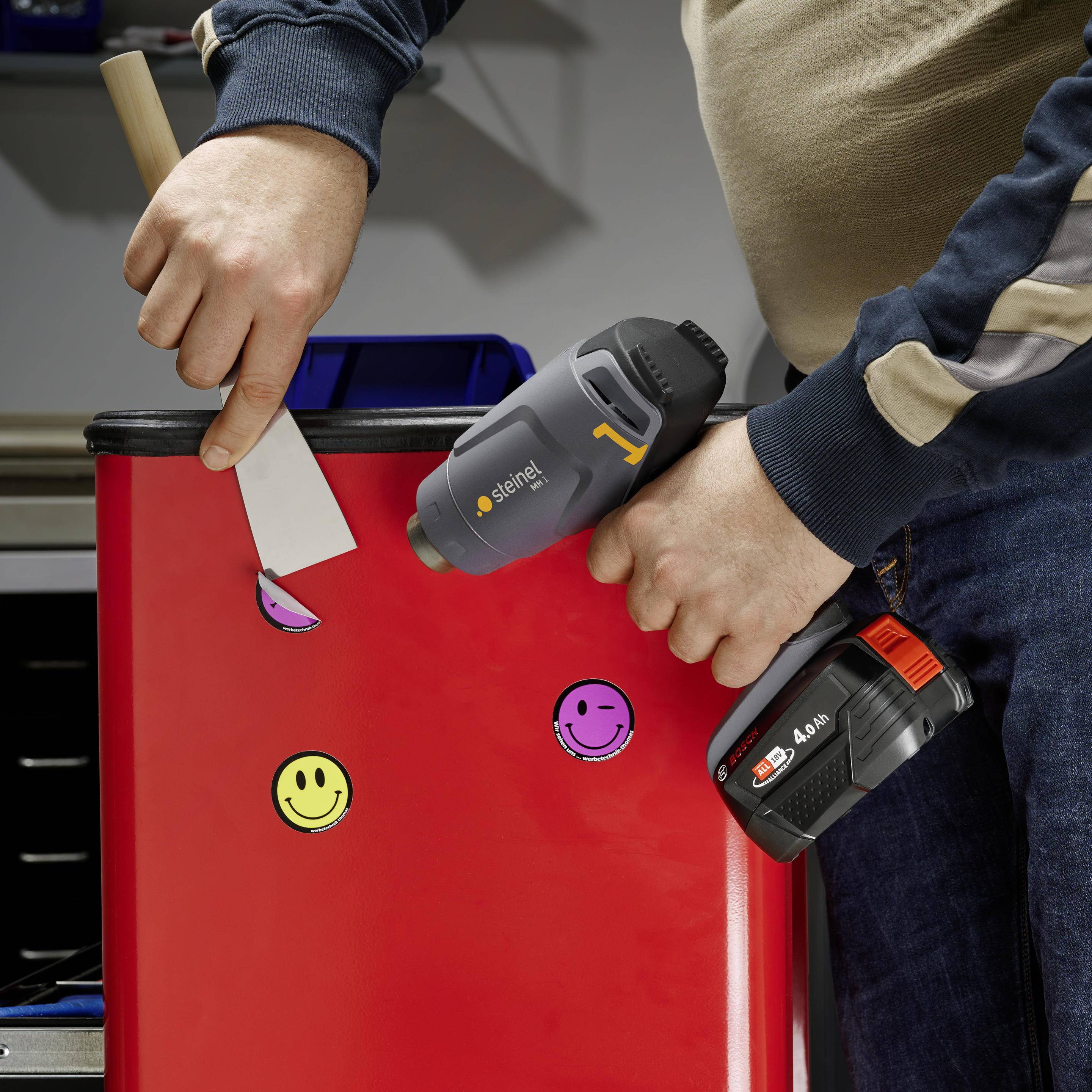 Eine Person klebt bunte Smileys auf eine rote Metalloberfläche. In der einen Hand hält sie ein Heißluftgebläse, in der anderen einen Spachtel.