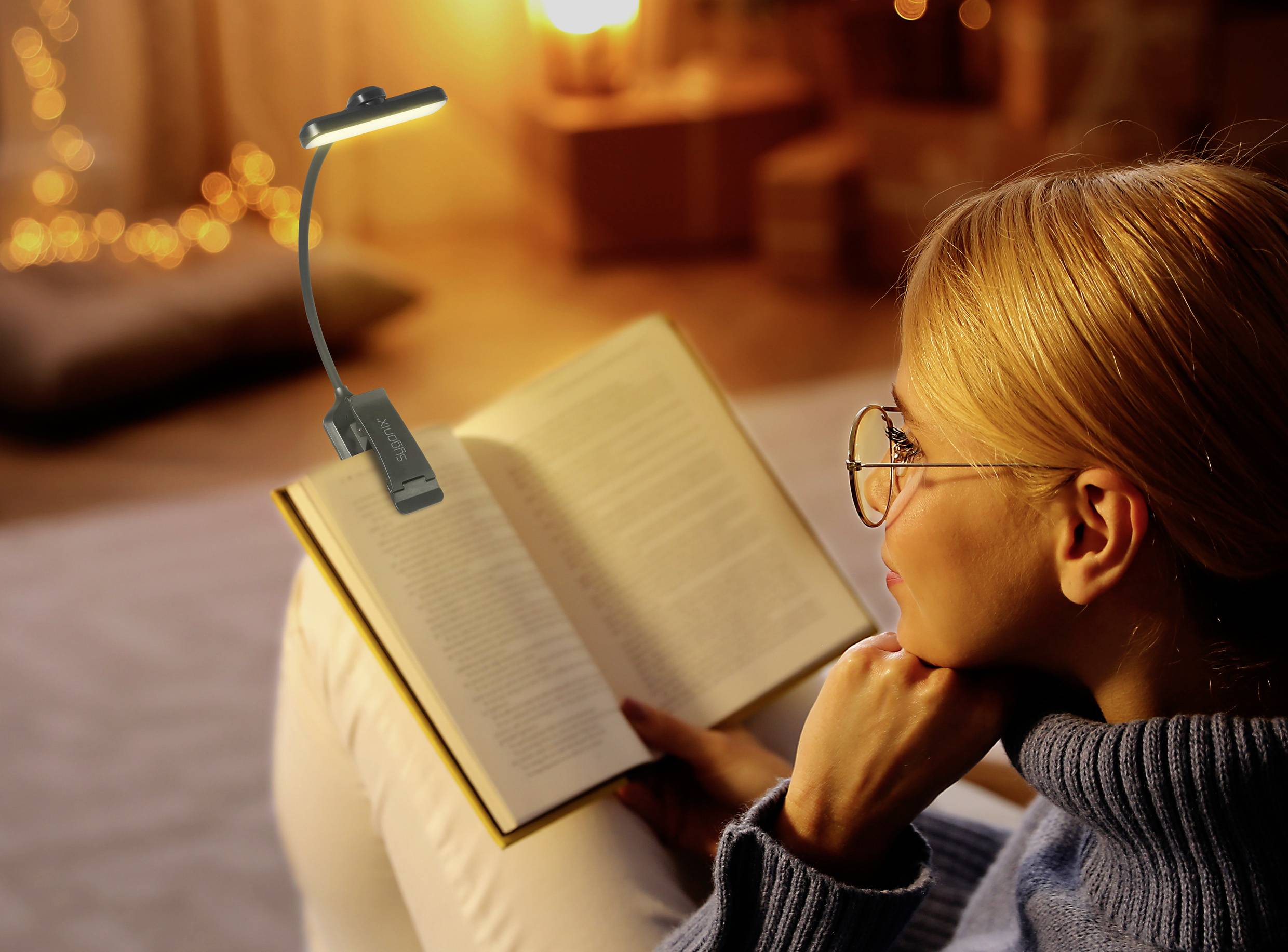 Eine Frau sitzt auf einem Sofa und liest ein Buch unter einer Leselampe. Es herrscht eine gemütliche, warme Atmosphäre im Raum.