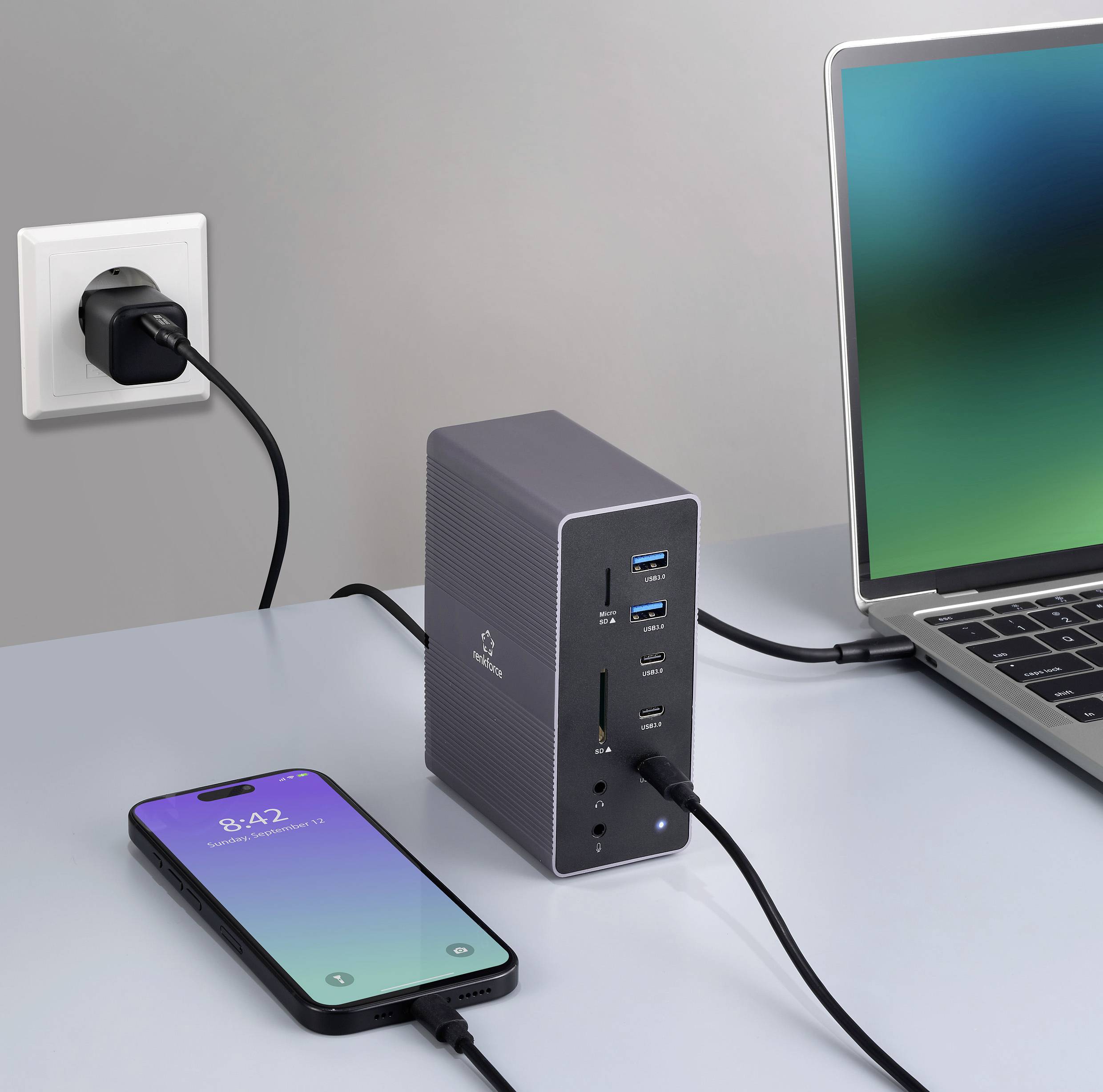 Ein Laptop ist über einen Hub mit mehreren Anschlüssen verbunden; ein Smartphone liegt daneben und lädt über das Netzteil an der Wand.