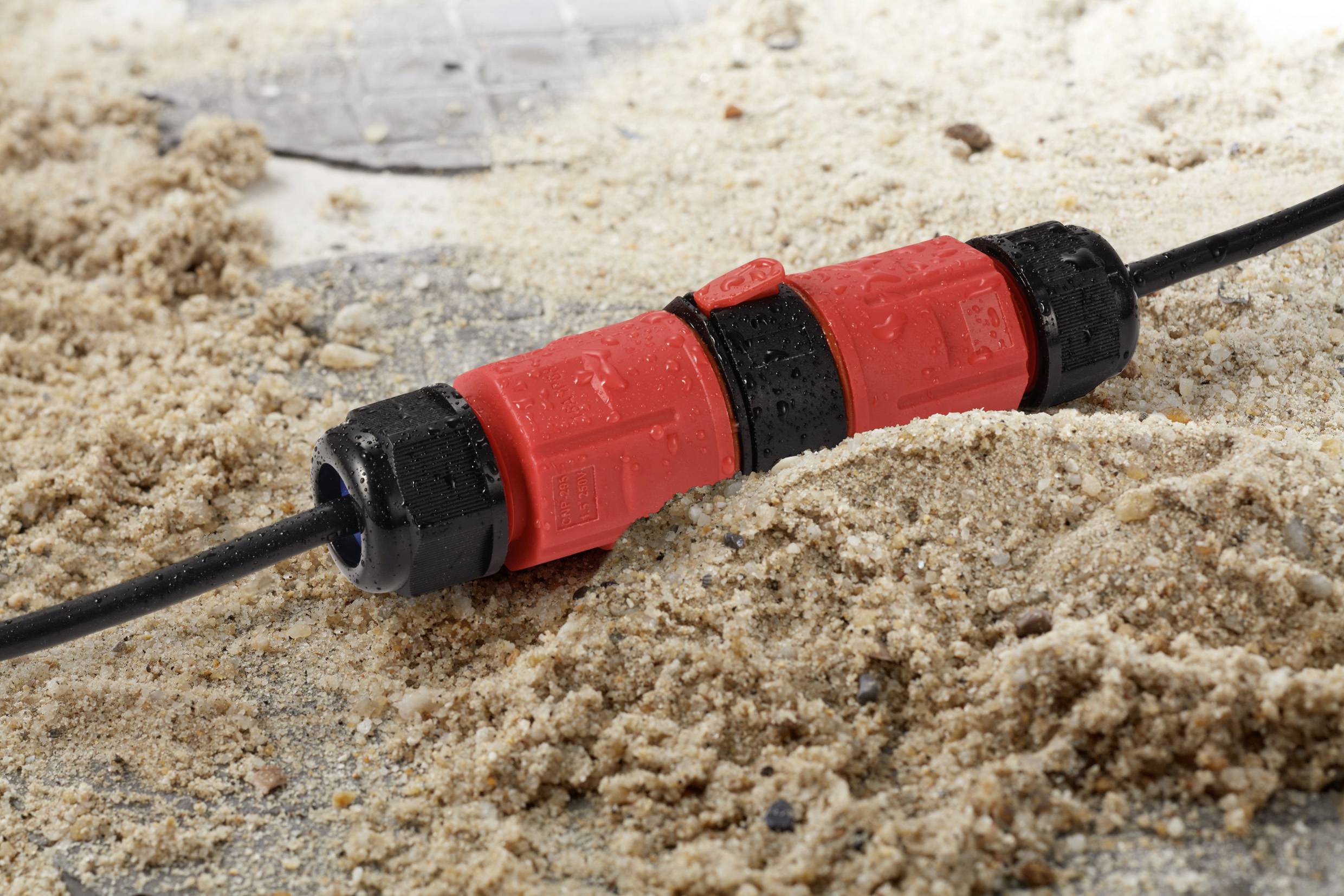 Schwarzes Kabel mit rotem, wasserdichtem Steckverbinder liegt teilweise im Sand. Der Verbinder zeigt Wassertropfen und Sandkörner.