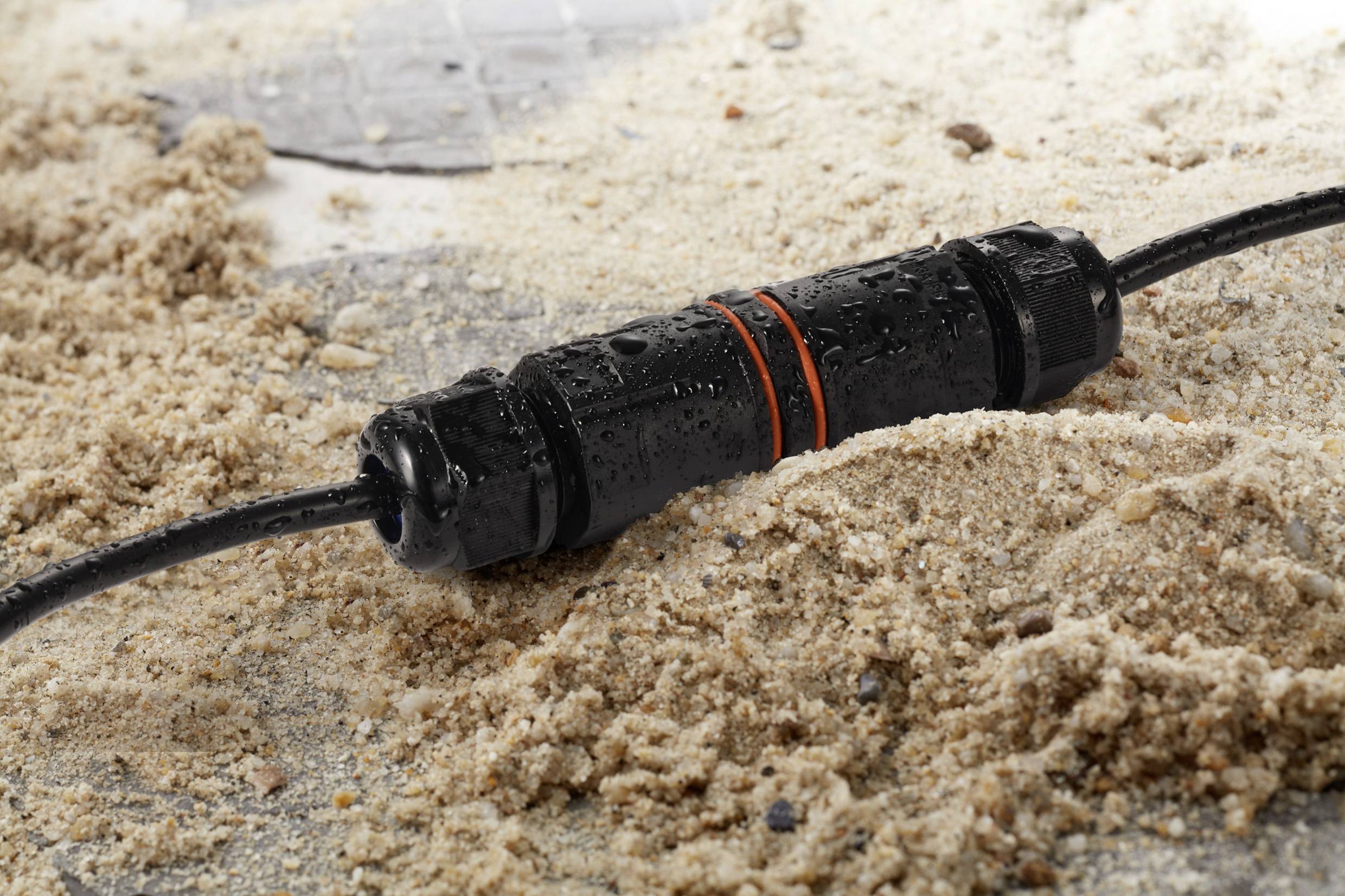 Ein wasserdichter Kabelverbinder liegt im Sand. Der Verbinder ist schwarz mit einer orangefarbenen Markierung und hat Wassertropfen darauf.