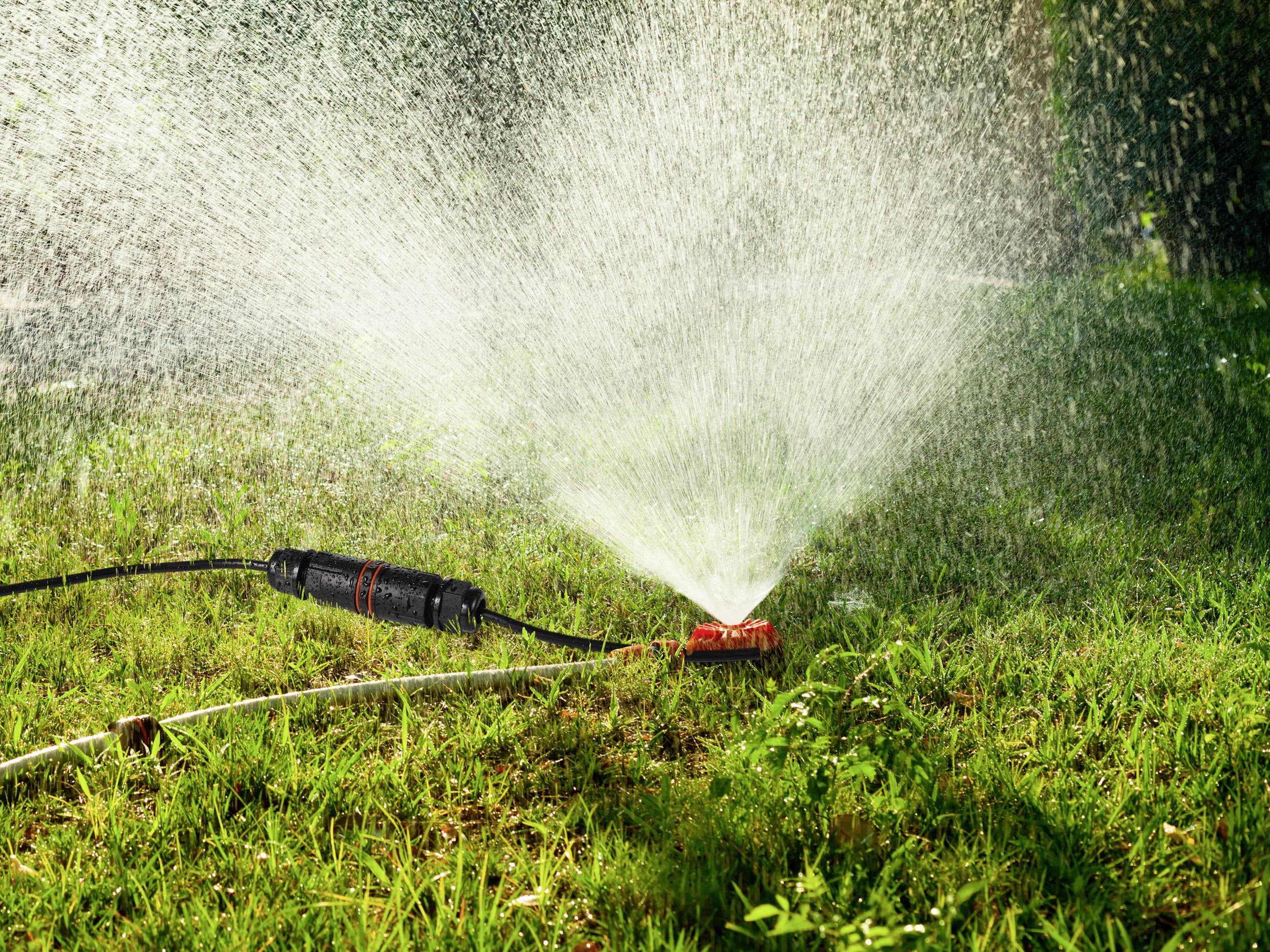 Ein Wasserregner bewässert grünes Gras bei Sonnenlicht, während feiner Wassernebel in der Luft sichtbar ist.