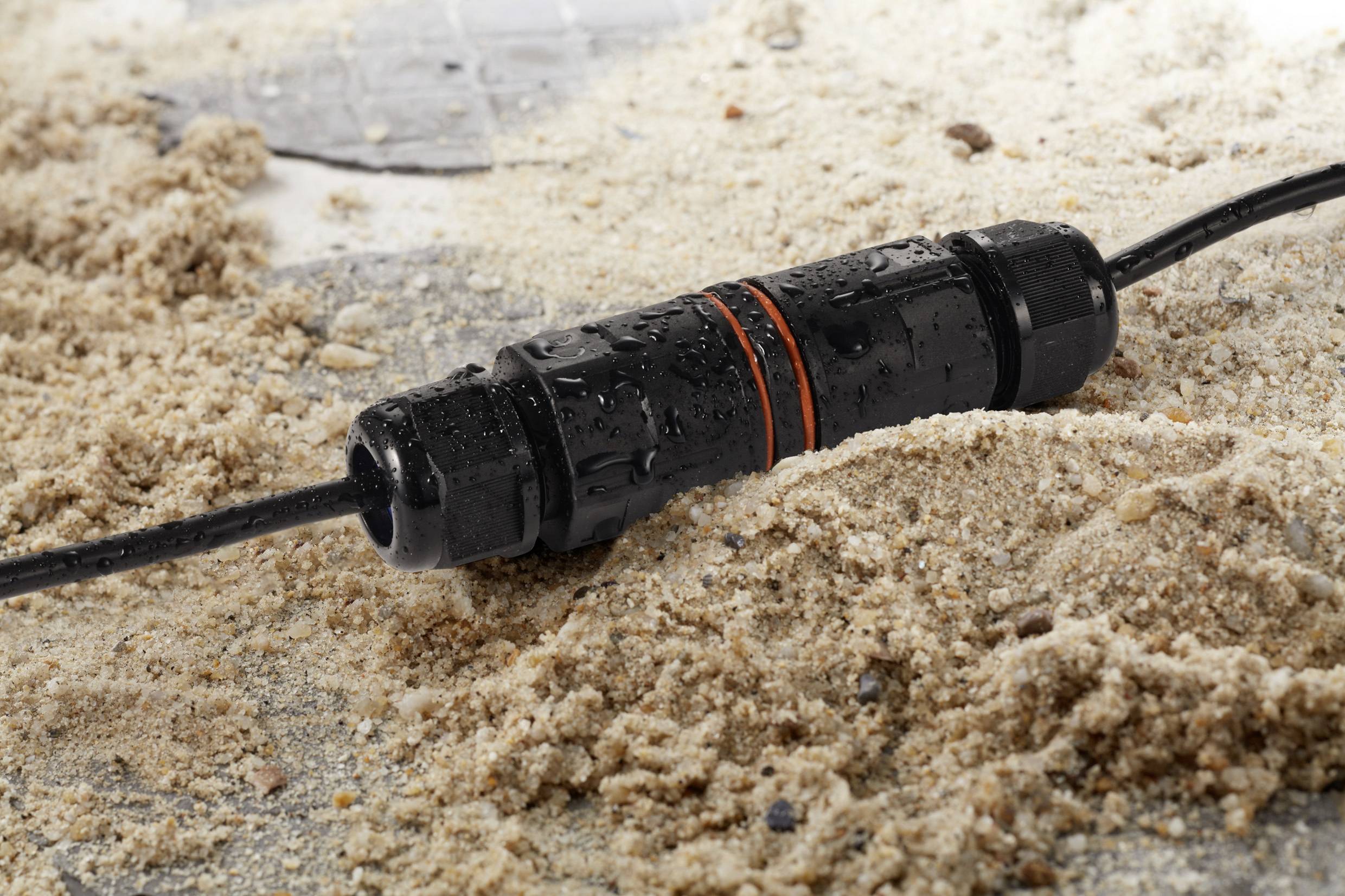 Ein wasserdichter Kabelverbinder liegt auf sandigem Untergrund. Das Kabel ist schwarz mit zwei orangen Ringen und zeigt Wassertröpfchen.