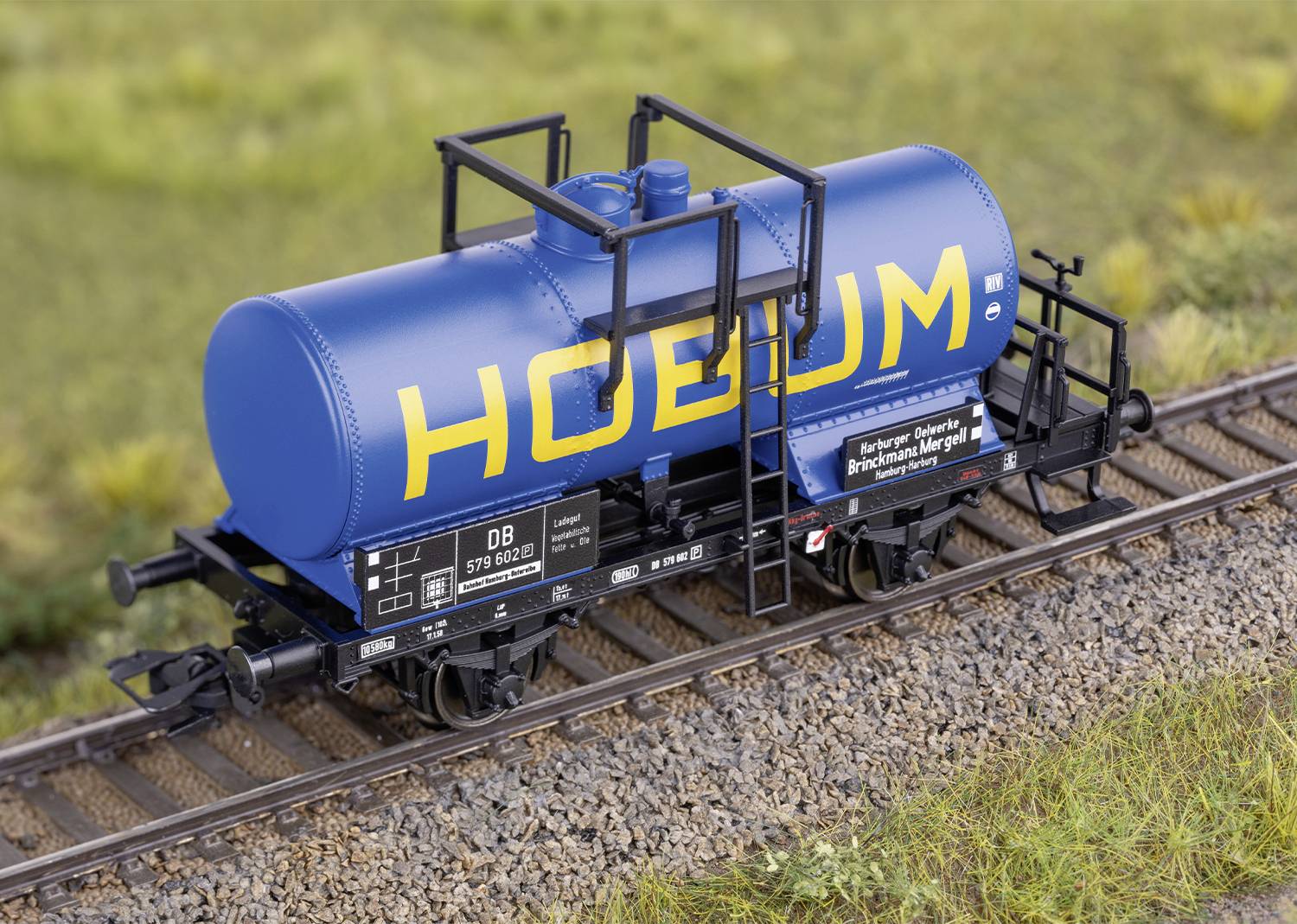 Ein blauer Kesselwagen auf einer Modelleisenbahnschiene. Auf dem Tank steht der Schriftzug 'HOEBUM' in gelber Farbe.