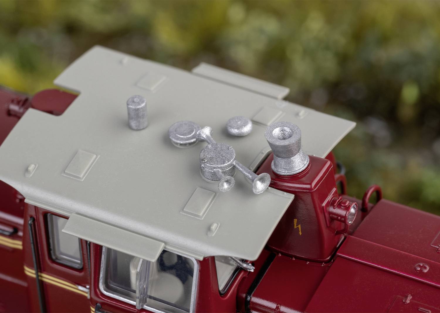 Miniaturmodell eines roten Fahrzeugs mit grauem Dach, auf dem kleine silberne Objekte platziert sind. Im Hintergrund ist unscharf Grün zu sehen.