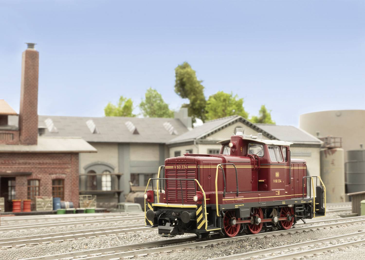 Eine Modell-Dampflokomotive in rot steht auf einem Gleis vor einem Miniatur-Industriegebäude mit Schornstein und Wassertank.