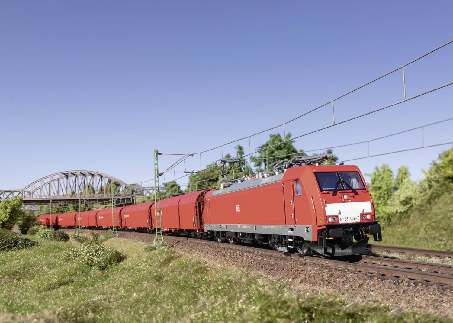 Ein roter Güterzug fährt tagsüber über eine Eisenbahnstrecke durch eine ländliche Gegend. Im Hintergrund ist eine Brücke zu sehen.