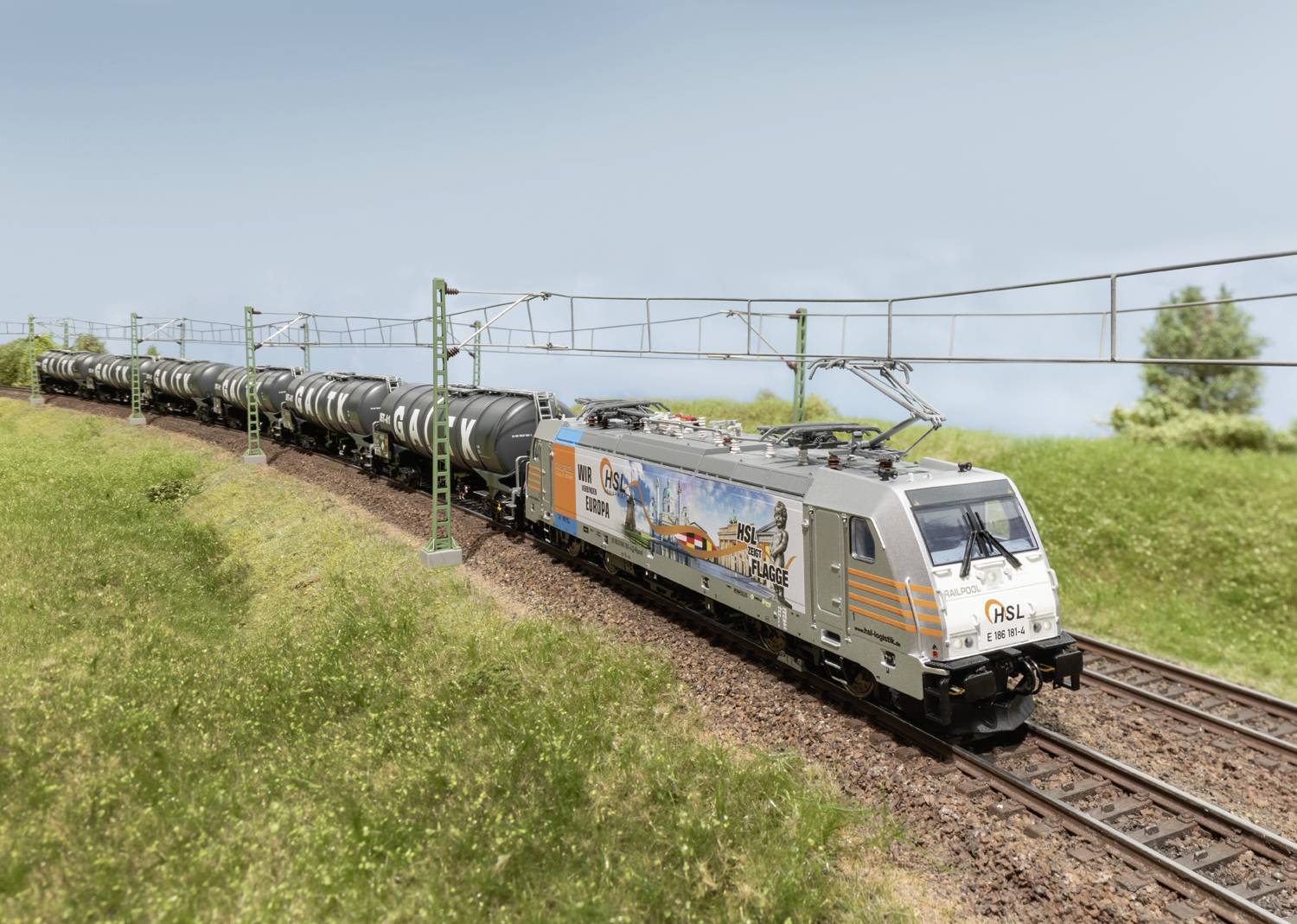 Ein Güterzug mit Tankwaggons fährt auf einer ländlichen Bahnstrecke. Auf der Lokomotive ist eine bunte Werbegrafik zu sehen.