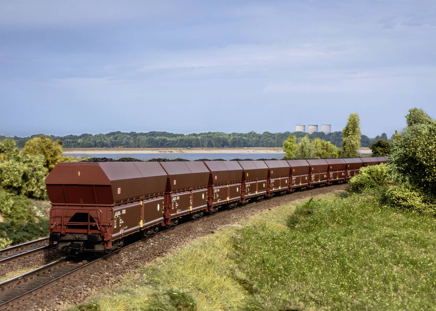 Ein langer Güterzug mit braunen Waggons fährt auf einer kurvigen Eisenbahnstrecke durch eine grüne Landschaft, im Hintergrund ein Kraftwerk.