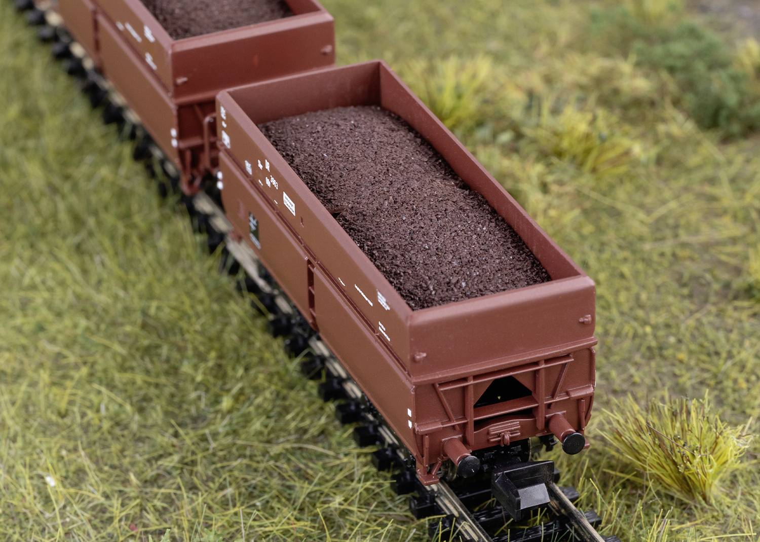 Ein Modellzug mit brauner Fracht, der über eine eisenbahnähnliche Miniaturlandschaft mit Gras fährt. Der Fokus liegt auf dem Zug.