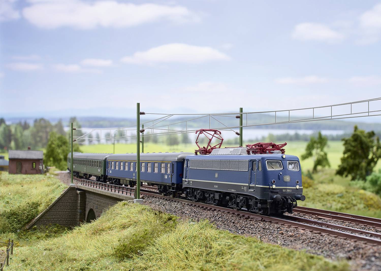 Ein blau-grauer Zug fährt über eine Brücke in einer ländlichen Landschaft. Hintergrund zeigt Felder und wolkigen Himmel.