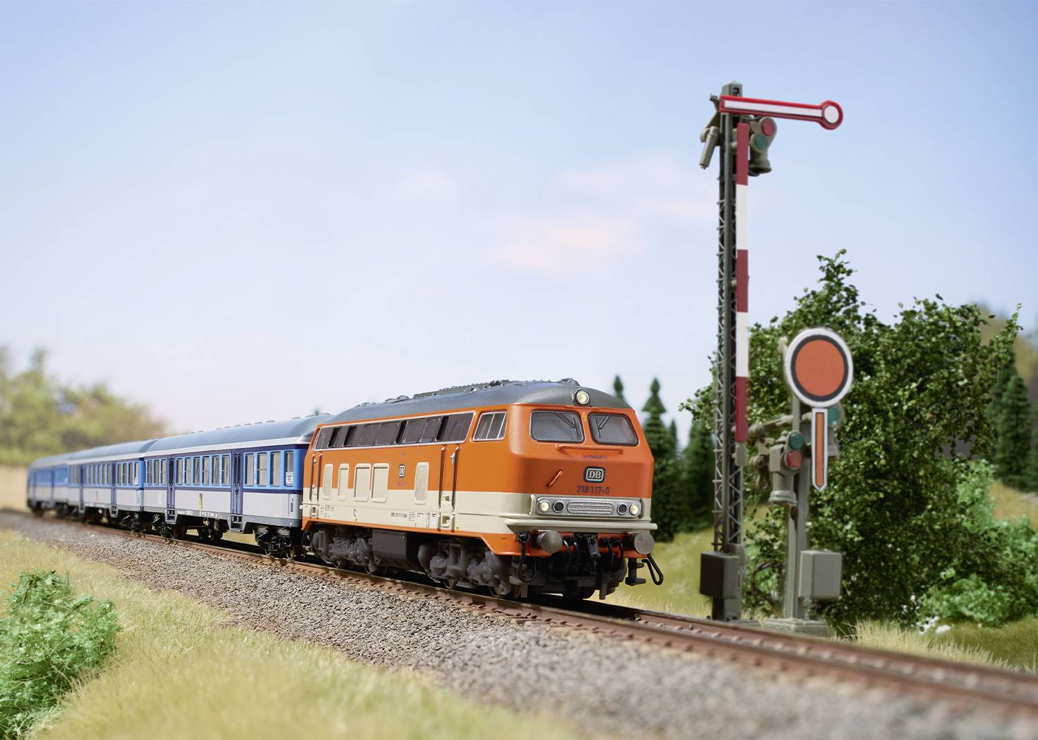 Ein orange-blauer Zug fährt auf einem Bahngleis durch eine grüne Landschaft. Ein mechanisches Signal steht auf Halt.