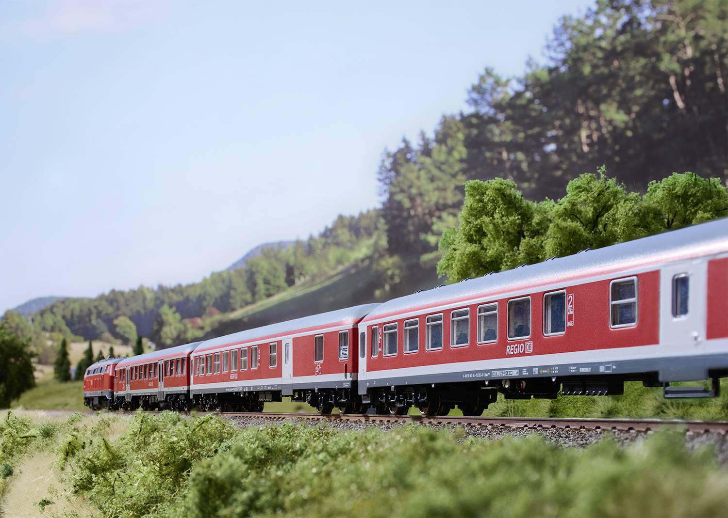 Ein roter Zug fährt durch eine ländliche Landschaft, umgeben von grünen Bäumen und Hügeln unter einem blauen Himmel.