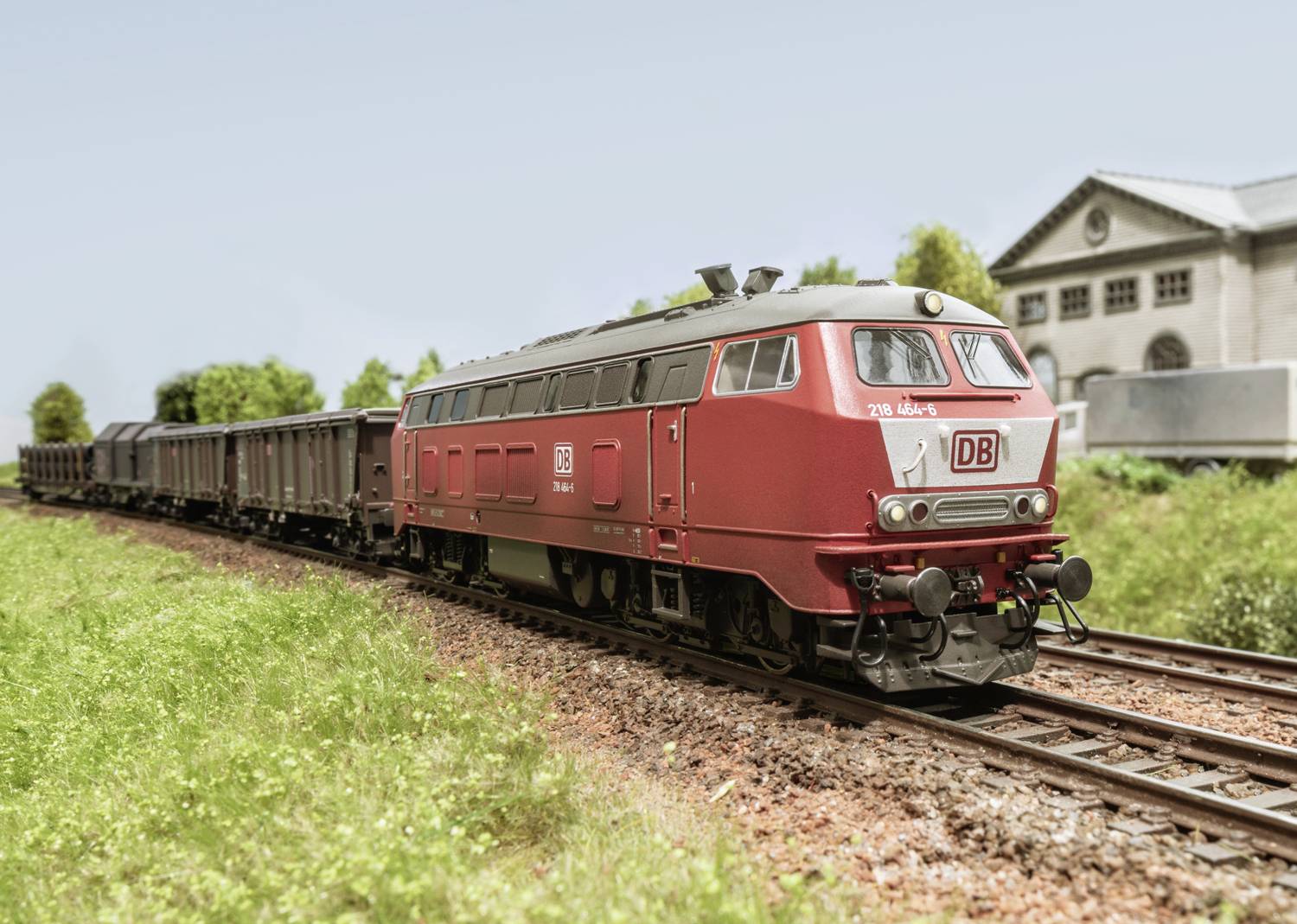 Ein roter Güterzug der Deutschen Bahn fährt auf einer grünen Wiese in ländlicher Umgebung. Im Hintergrund ein Gebäude.