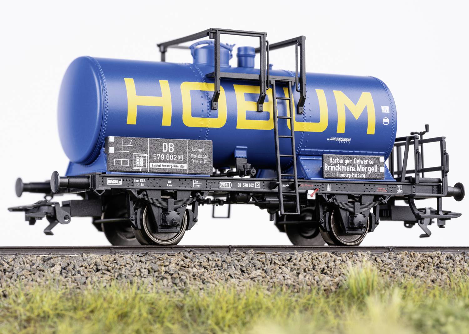 Ein blauer Modellzugkesselwagen auf Schienen mit der Aufschrift 'Hoelum' in gelb. Umgeben von Gras und Stein.