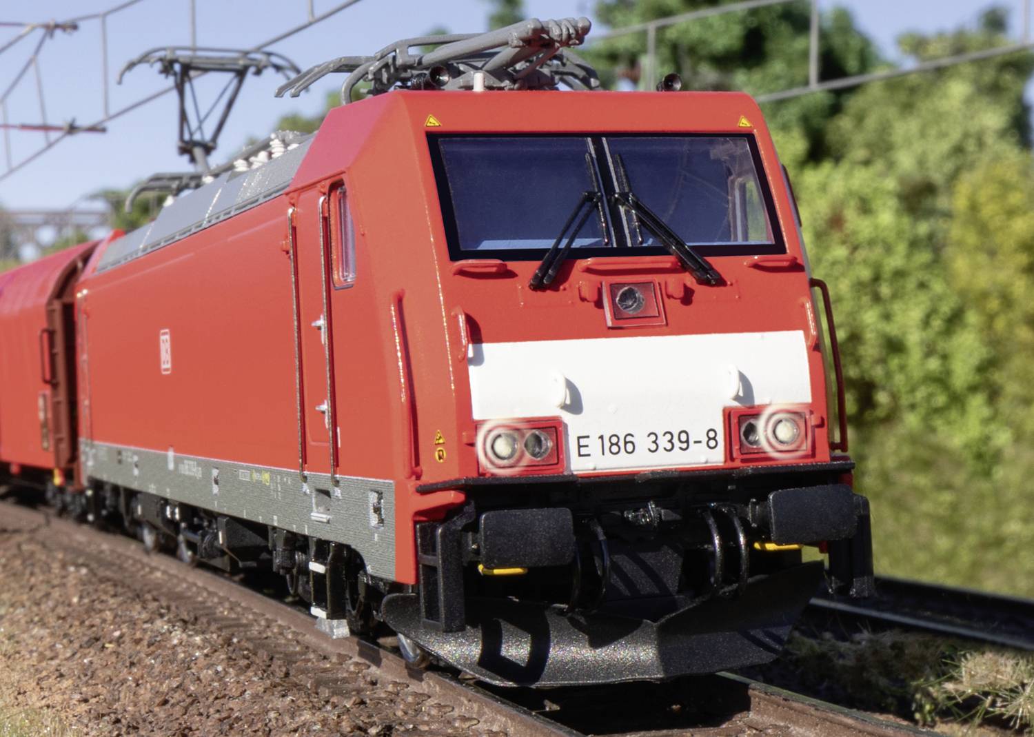 Eine rote Lokomotive fährt auf einem Gleis durch eine grüne Landschaft. Die Sonne scheint, und die Lokomotive hat die Nummer 'E 186 339-8'.