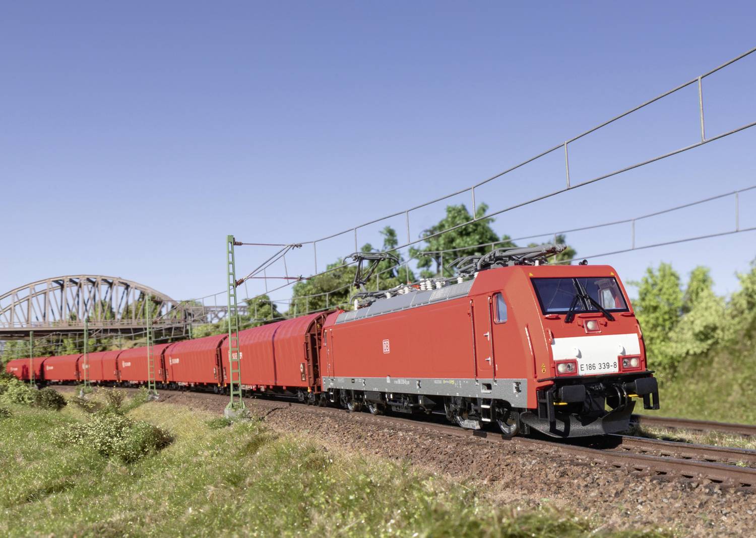 Ein roter Güterzug fährt bei klarem Himmel durch eine grüne Landschaft und passiert eine Eisenbahnbrücke im Hintergrund.